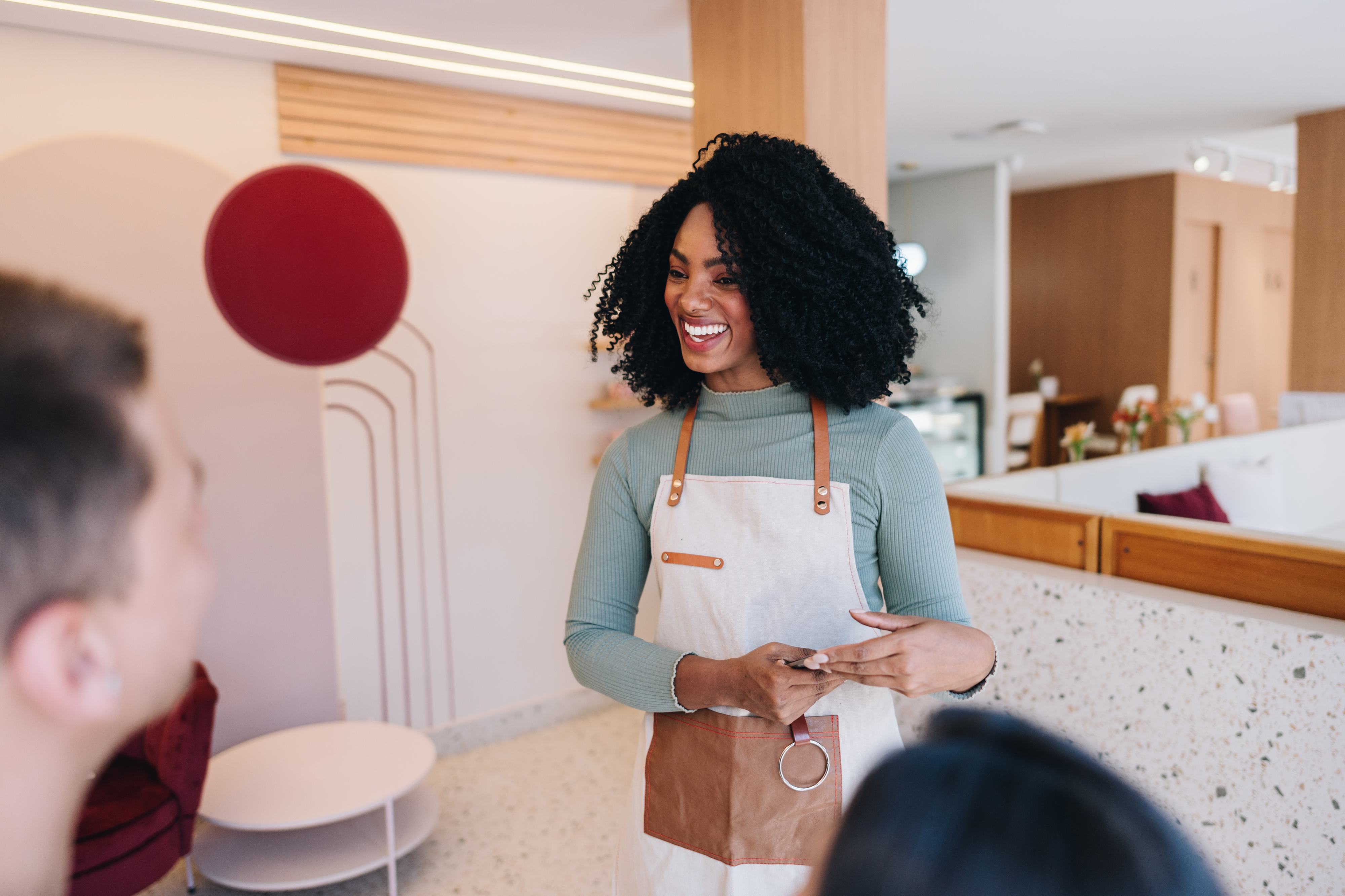 A woman with curly hair, wearing a green sweater and an apron, smiles while talking to two people in a modern room with a minimalist design