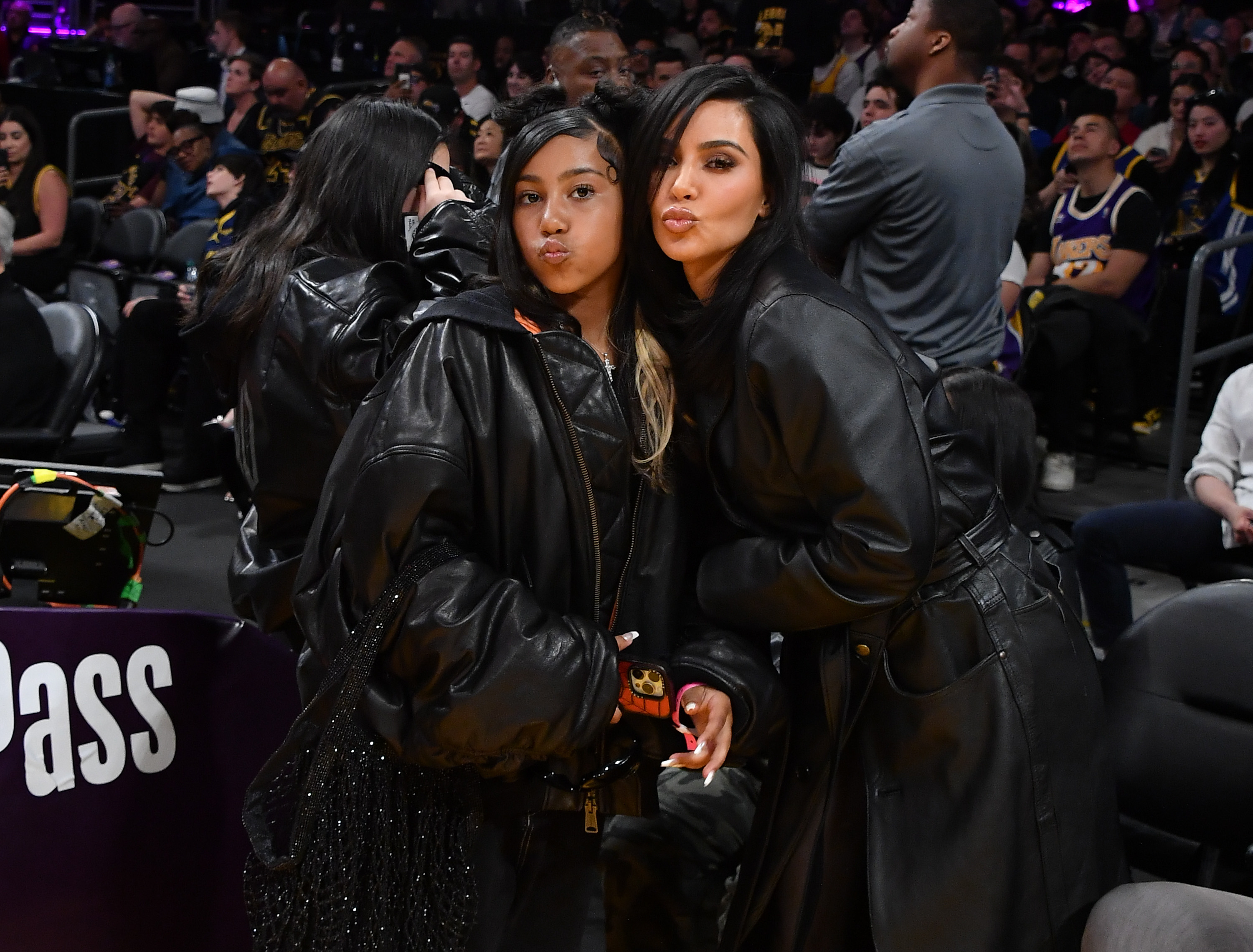 Kim Kardashian and her daughter, North West, both dressed in matching dark oversized leather jackets, posing at a basketball game with audience members in the background