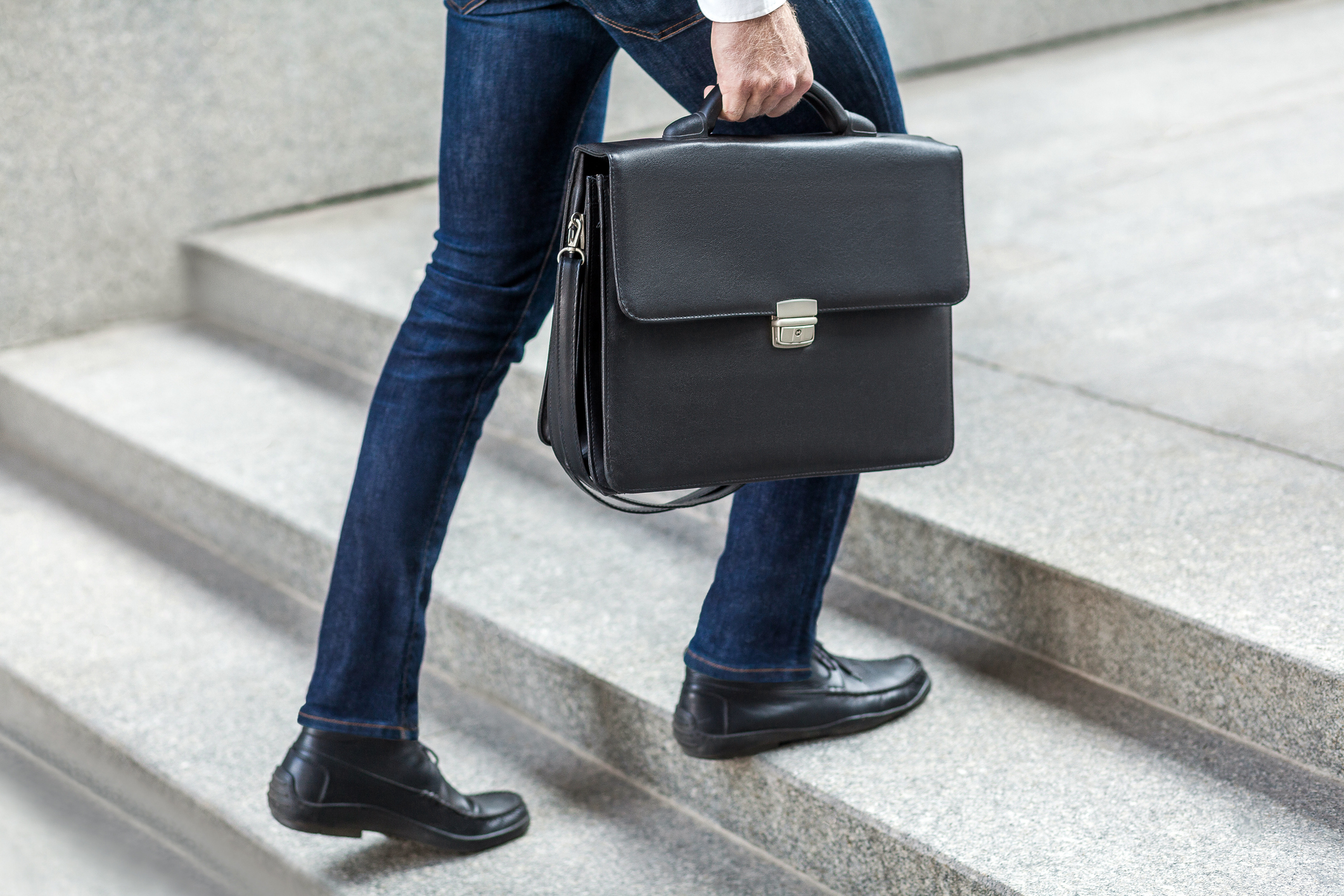 Person in jeans and dress shoes carrying a briefcase while walking up a staircase