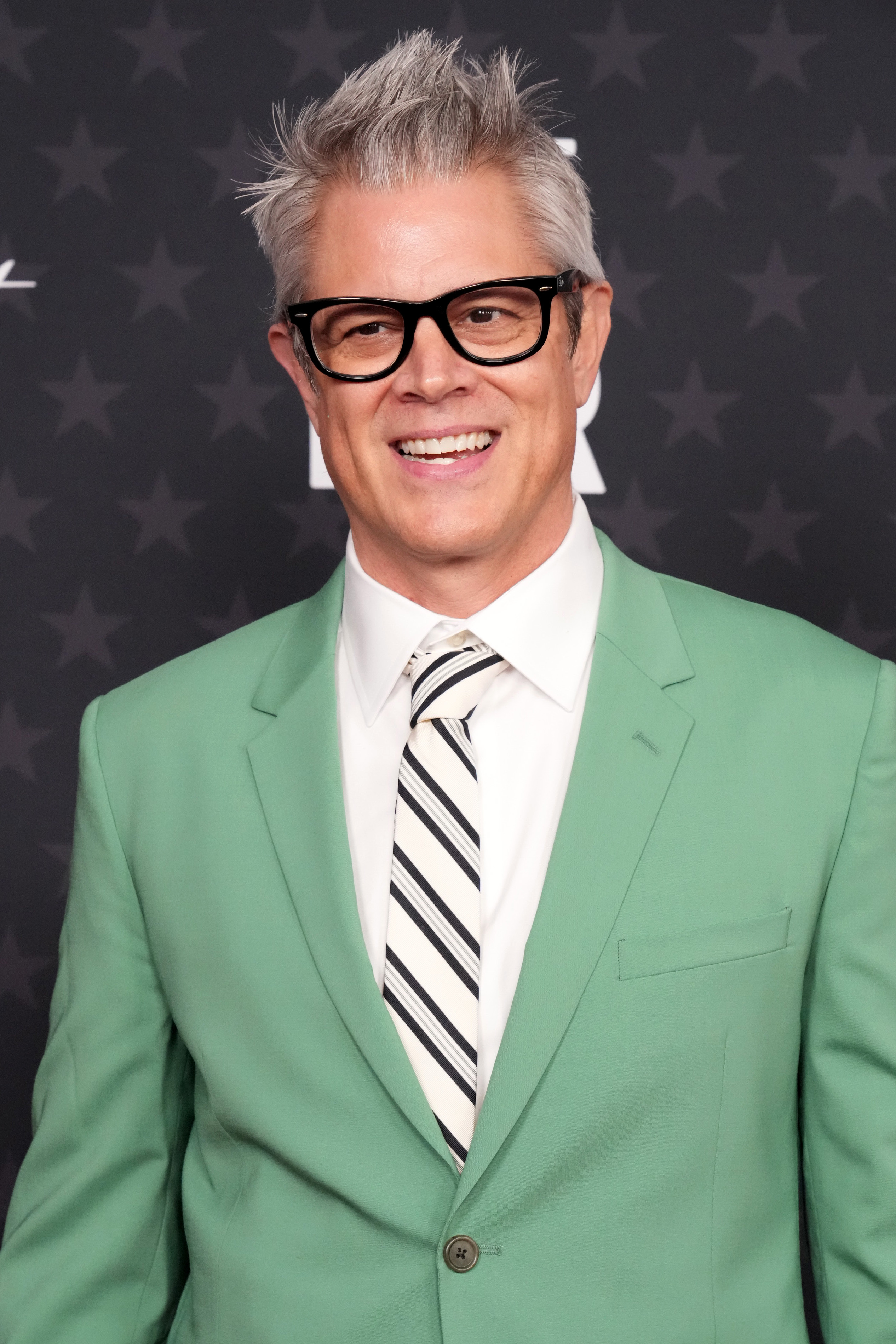 Johnny Knoxville in a light green suit and striped tie at a red carpet event, smiling at the camera