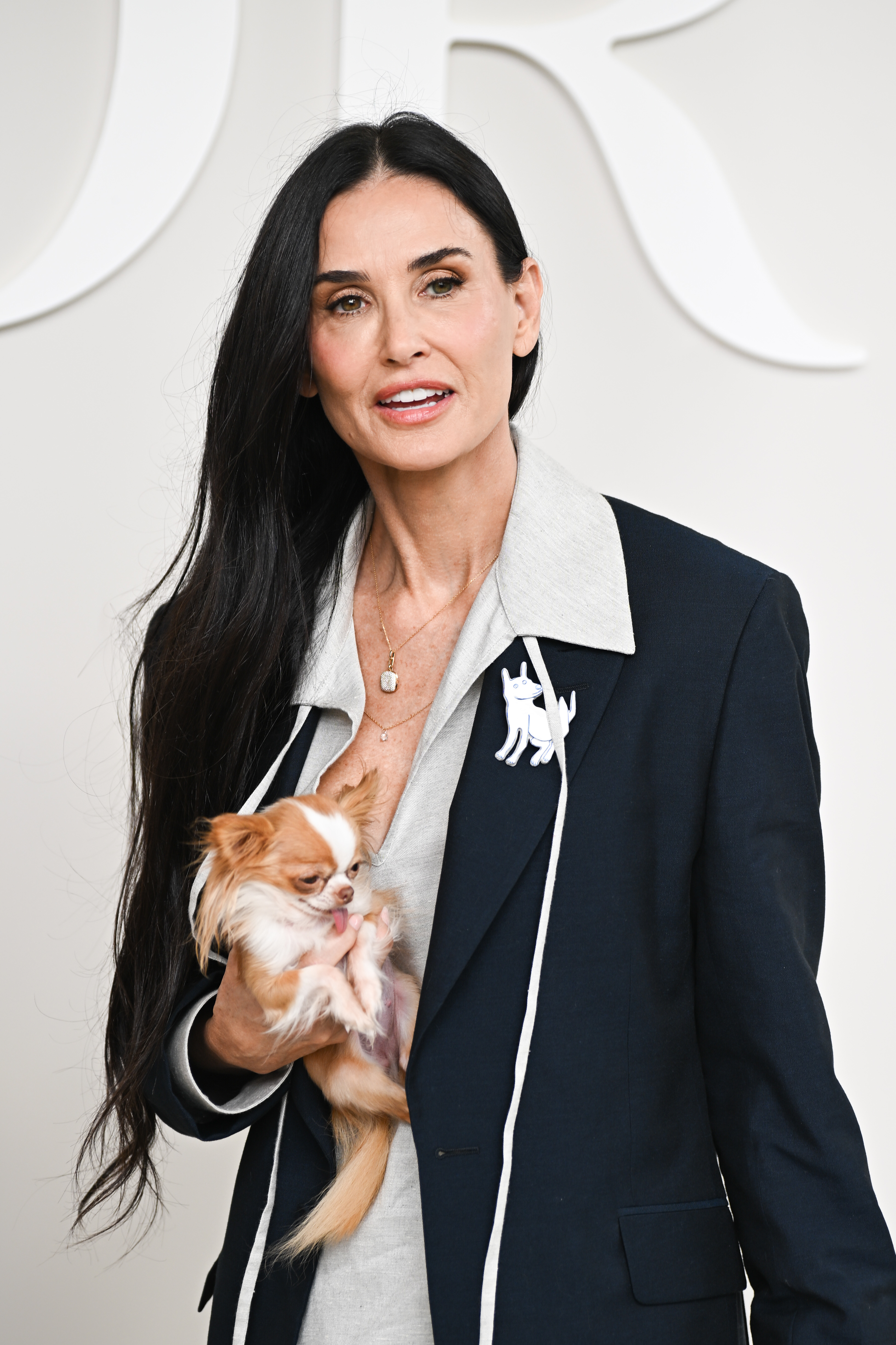 Demi Moore holding a small dog, dressed in a blazer over a shirt with a brooch, standing in front of a backdrop