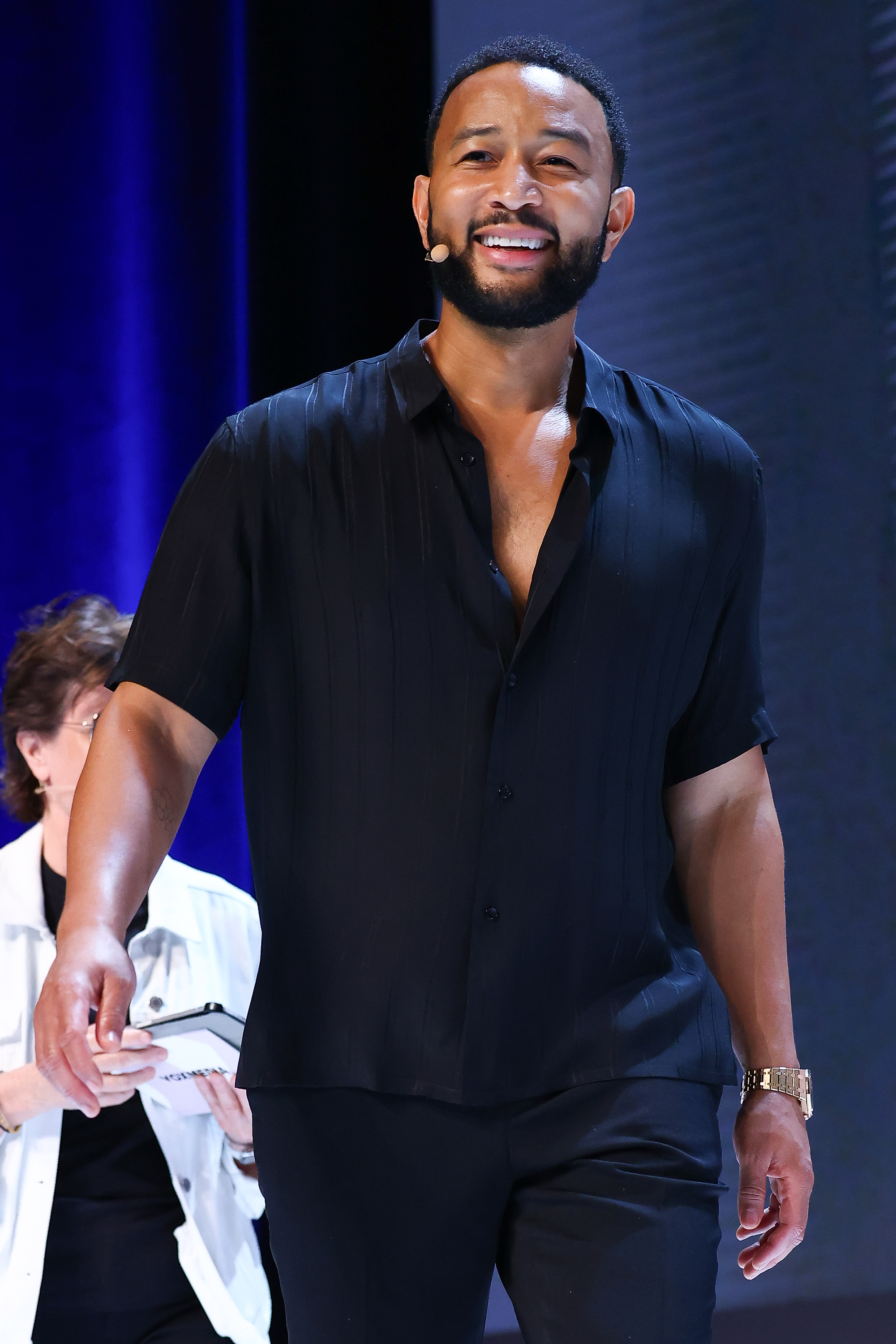 John Legend on stage, wearing a black shirt with a deep V-neck and matching pants. Another person in the background holds a notebook
