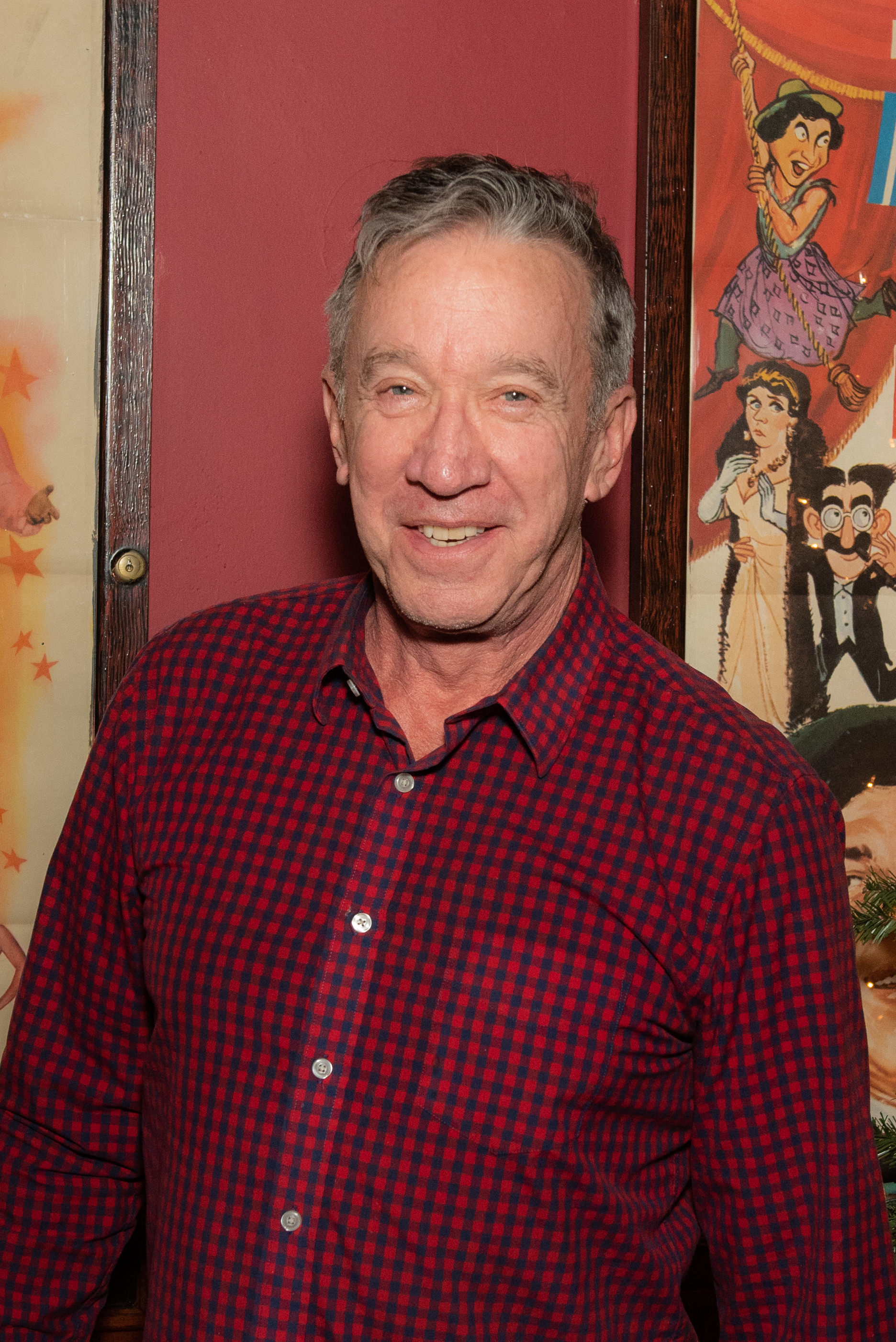 A man in a red and black checkered shirt smiling in front of a colorful poster with cartoon characters