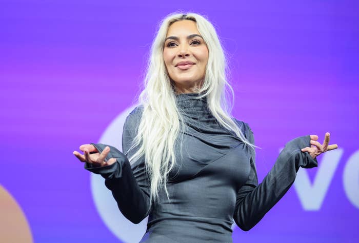 Kim Kardashian, wearing a fitted, long-sleeve dress, gestures with both hands while standing onstage