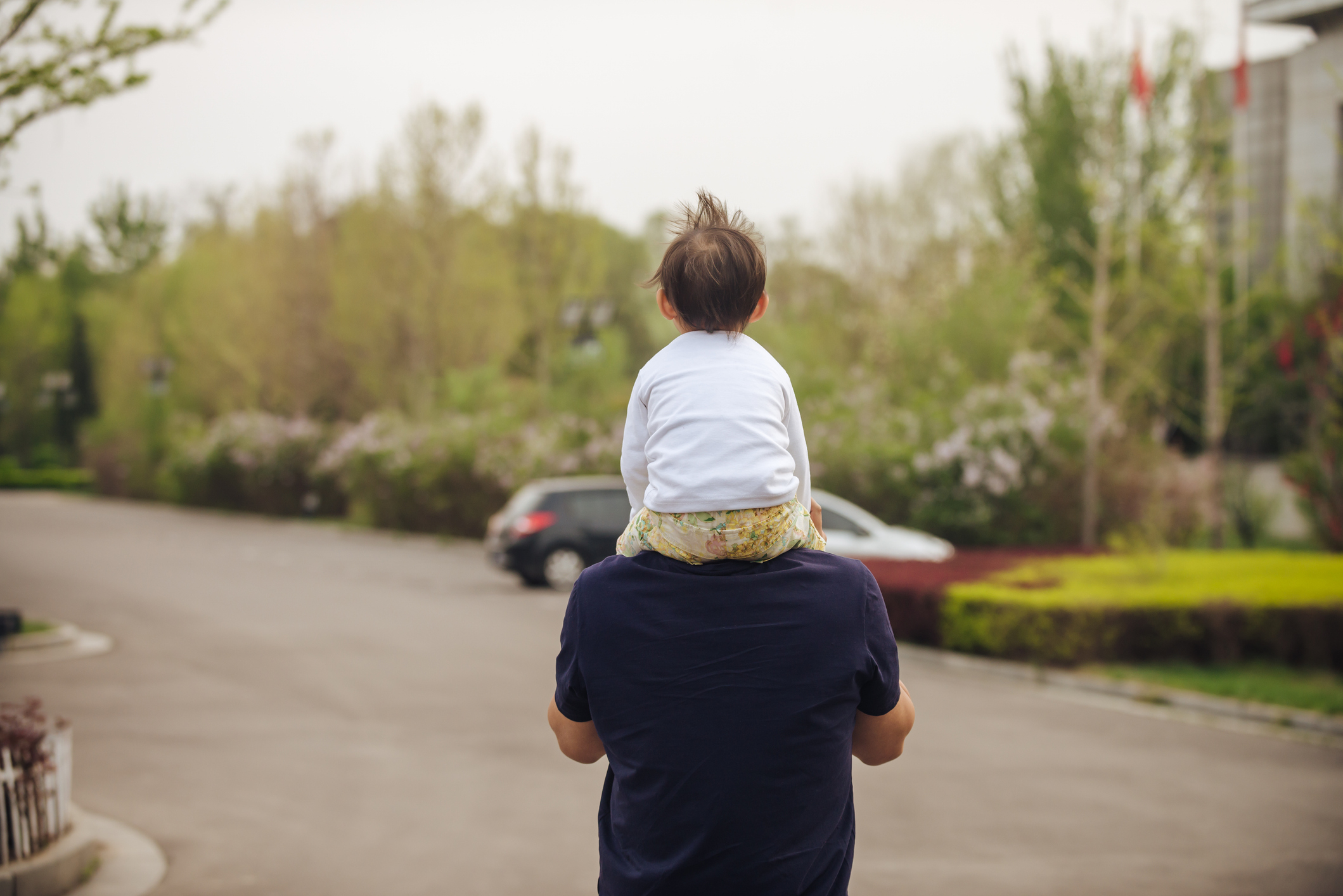 A person carries a small child on their shoulders while walking down a quiet, tree-lined street. Both are facing away from the camera