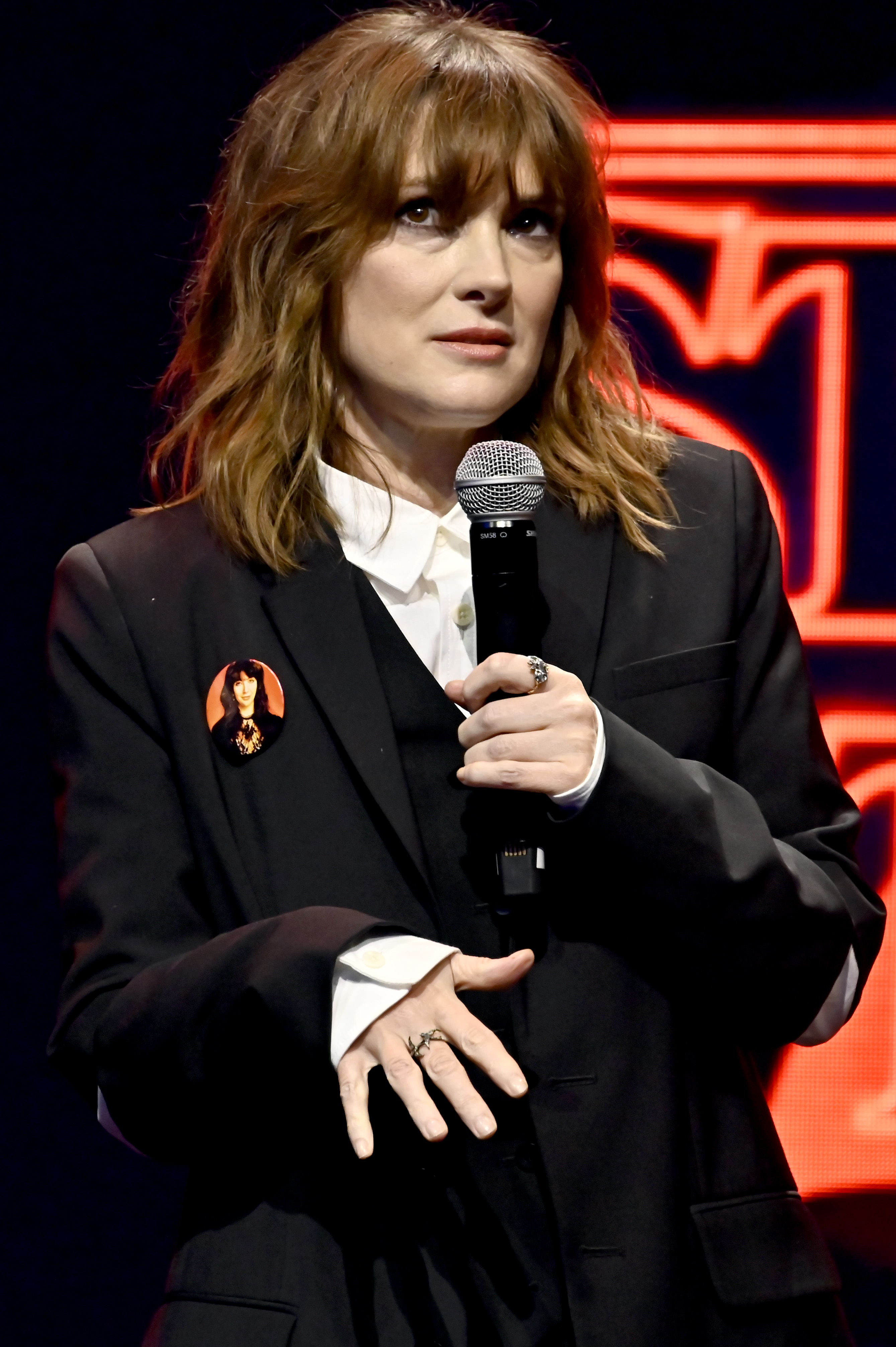 Winona Ryder speaks on stage holding a microphone, wearing a tailored black suit with a button featuring a woman's face