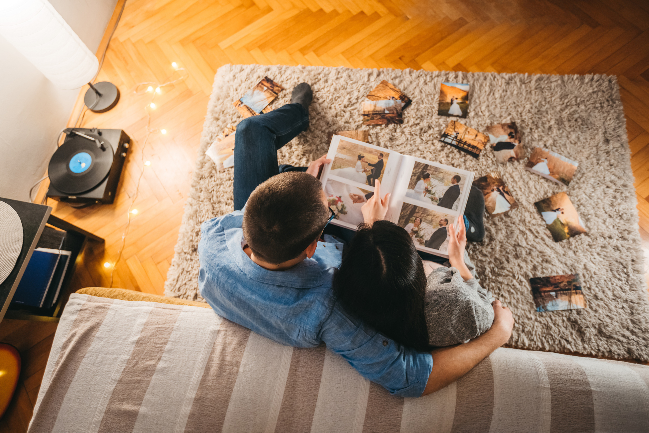 A couple is sitting on a couch, looking at a photo album together. There are several photos spread around them on the carpet, suggesting shared memories