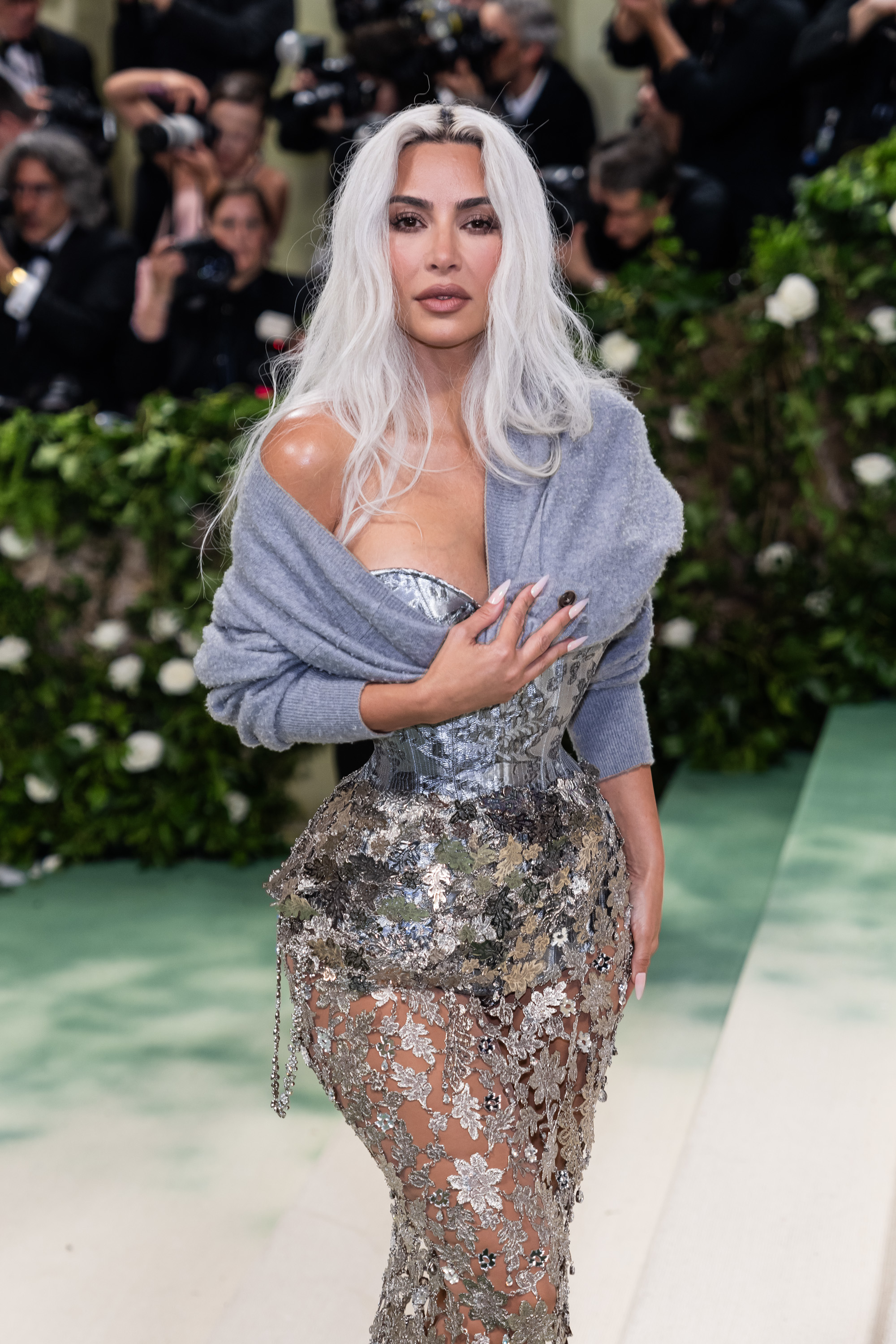 Kim Kardashian in a sparkling, form-fitting metallic dress with floral details and a shawl, posing on a green carpet at an event