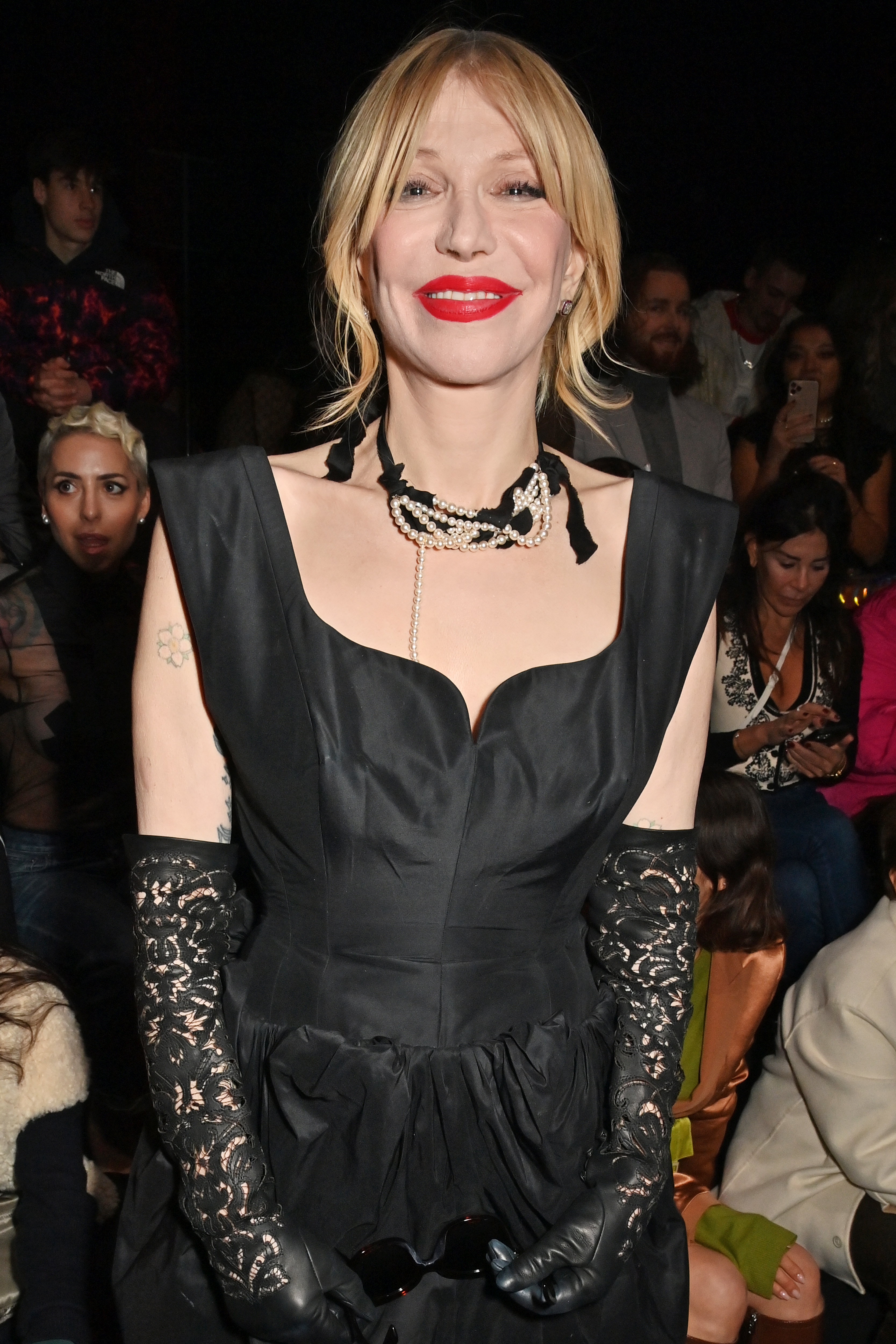 Courtney Love wearing a black dress with intricate gloves and pearl necklace at a public event