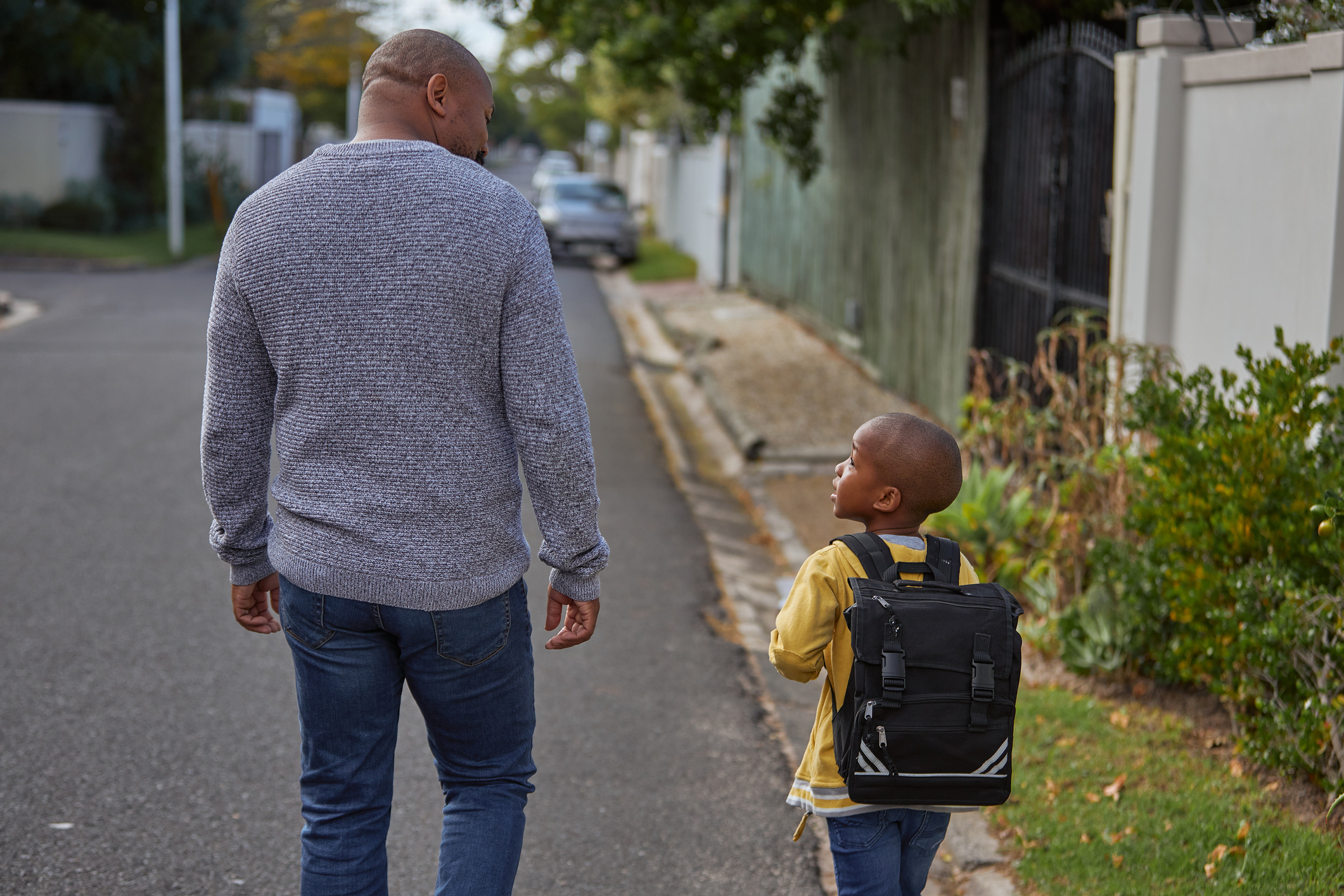 A man and a young boy are walking on a sidewalk. The man is looking down at the boy, who is wearing a backpack. They appear to be having a conversation
