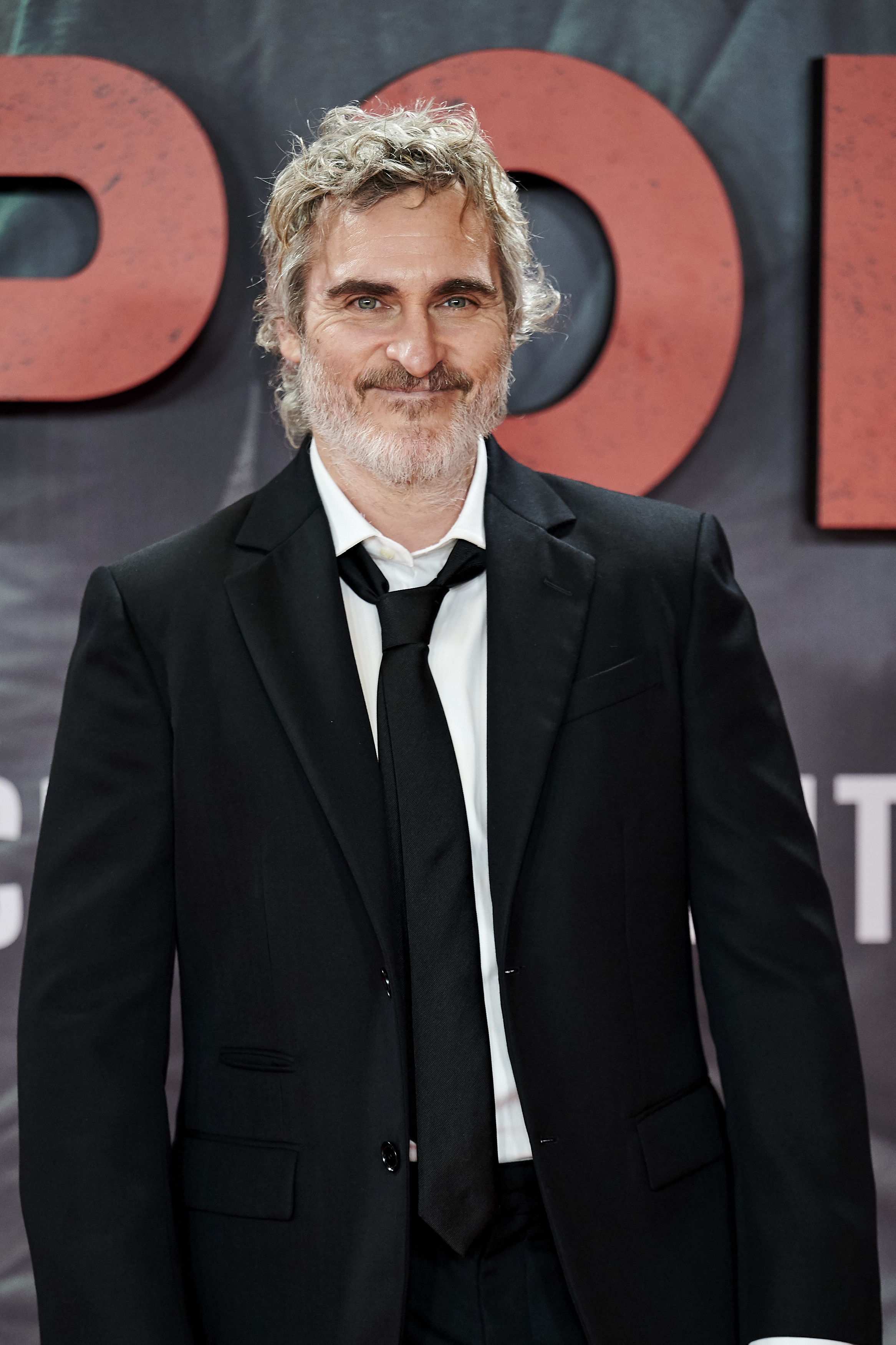 Joaquin Phoenix on the red carpet, wearing a black suit with a white dress shirt and black tie, smiling