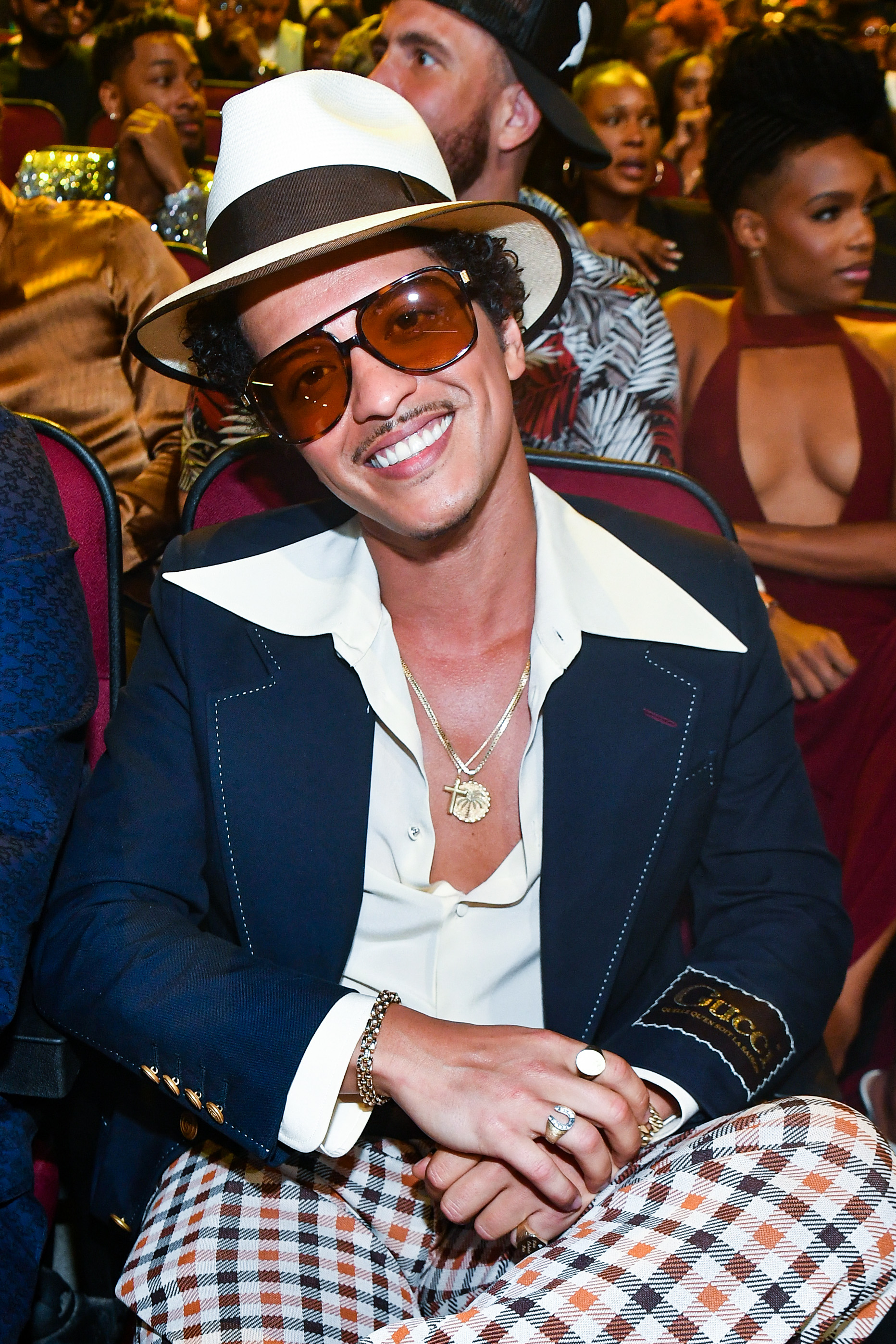 Bruno Mars smiling and seated at an event, wearing a fedora, aviator sunglasses, a navy blazer, a white shirt with a wide collar, and checkered pants