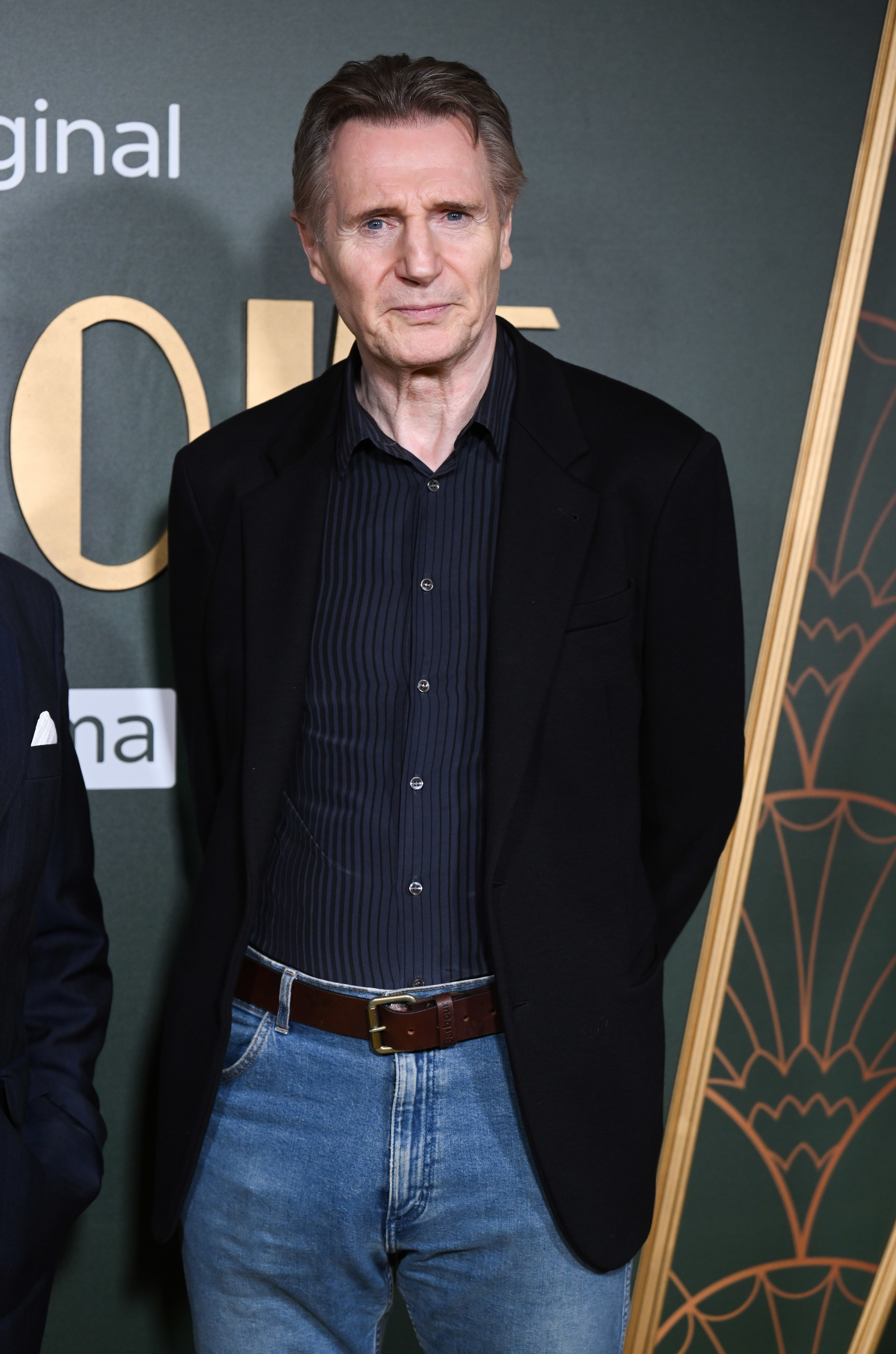 Liam Neeson stands in front of a movie premiere backdrop wearing a black blazer over a striped shirt and blue jeans