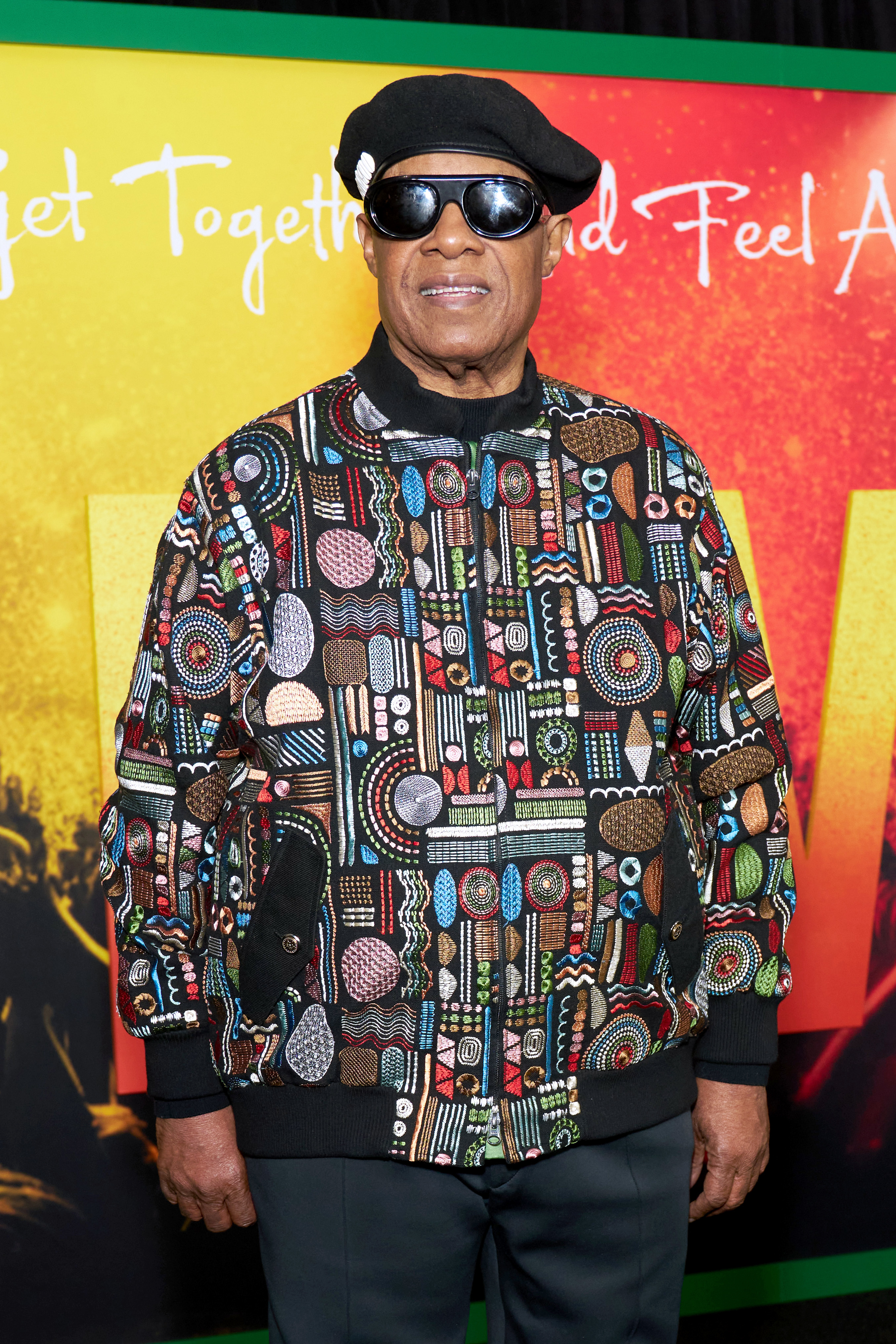 Stevie Wonder on the red carpet wearing a patterned jacket and a black hat