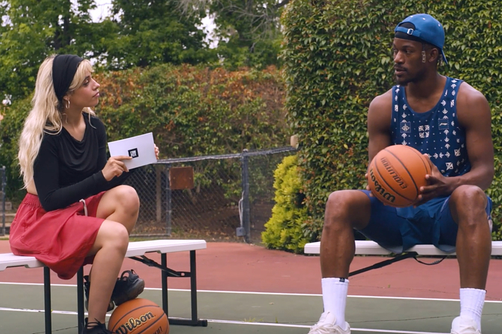 Camila Cabello holding a card with a microphone interviews Jimmy Butler in a sleeveless jersey and cap. Both hold basketballs while sitting on benches on an outdoor court