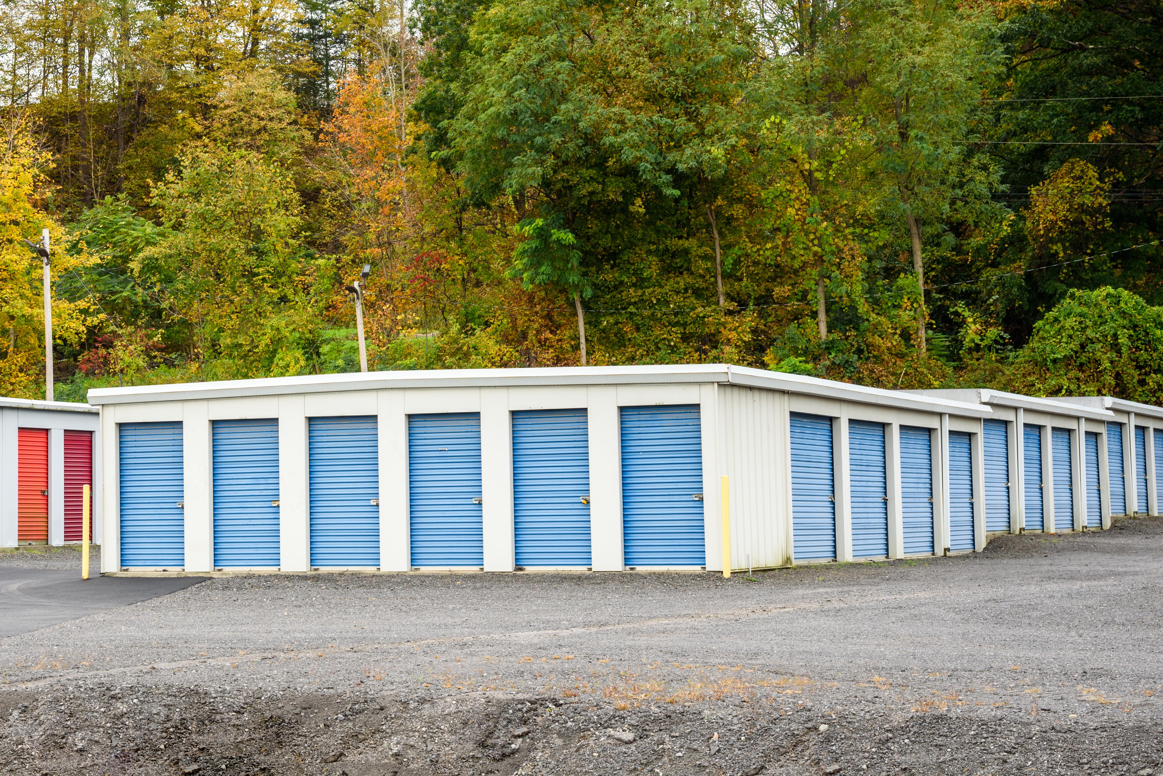 A row of storage units with blue doors located in a gravel area, surrounded by trees and foliage in the background. No people are in the image