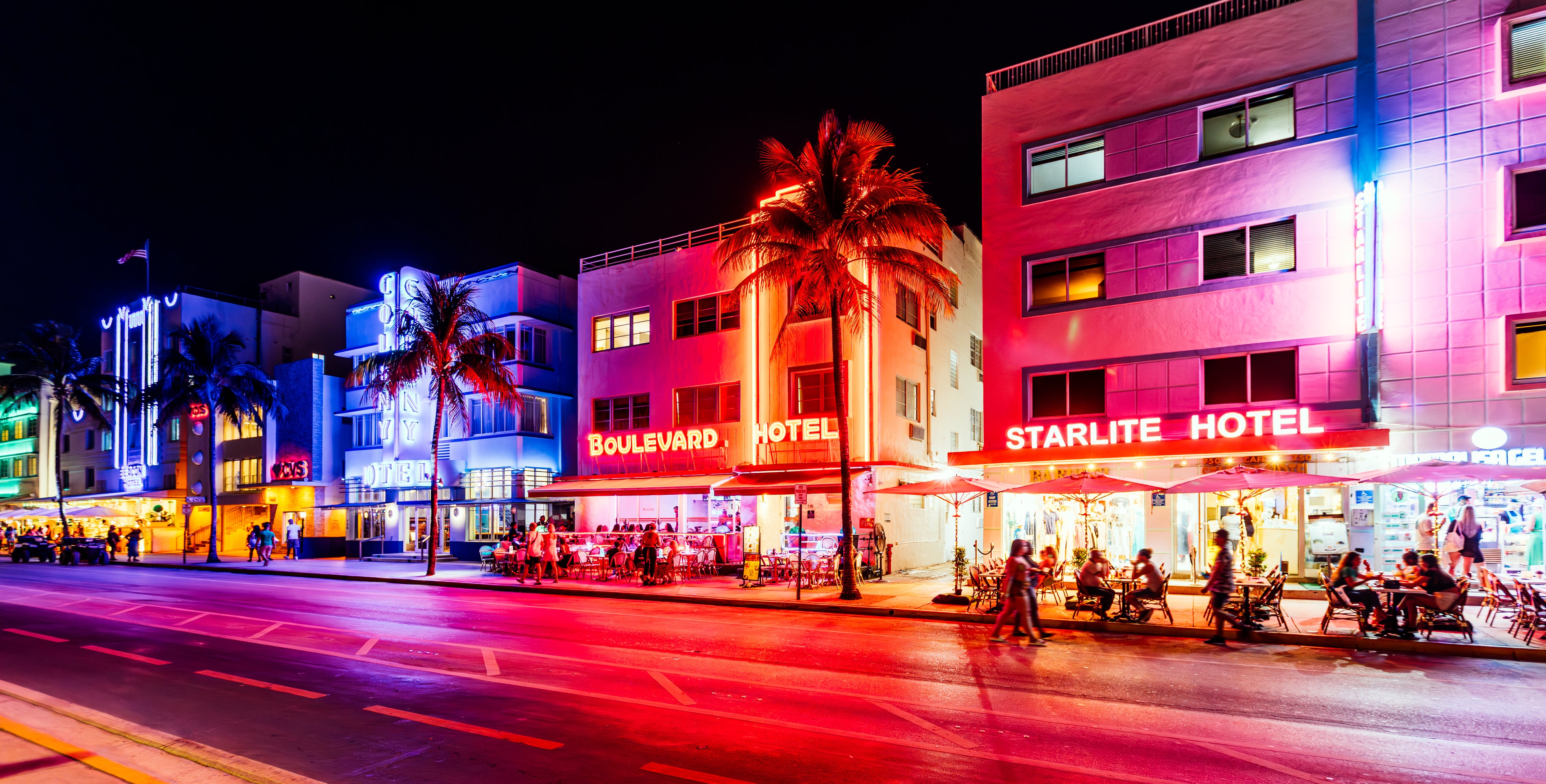 A vibrant night scene of Ocean Drive in Miami Beach showing neon-lit hotels, including Boulevard Hotel and Starlite Hotel, with people dining outside