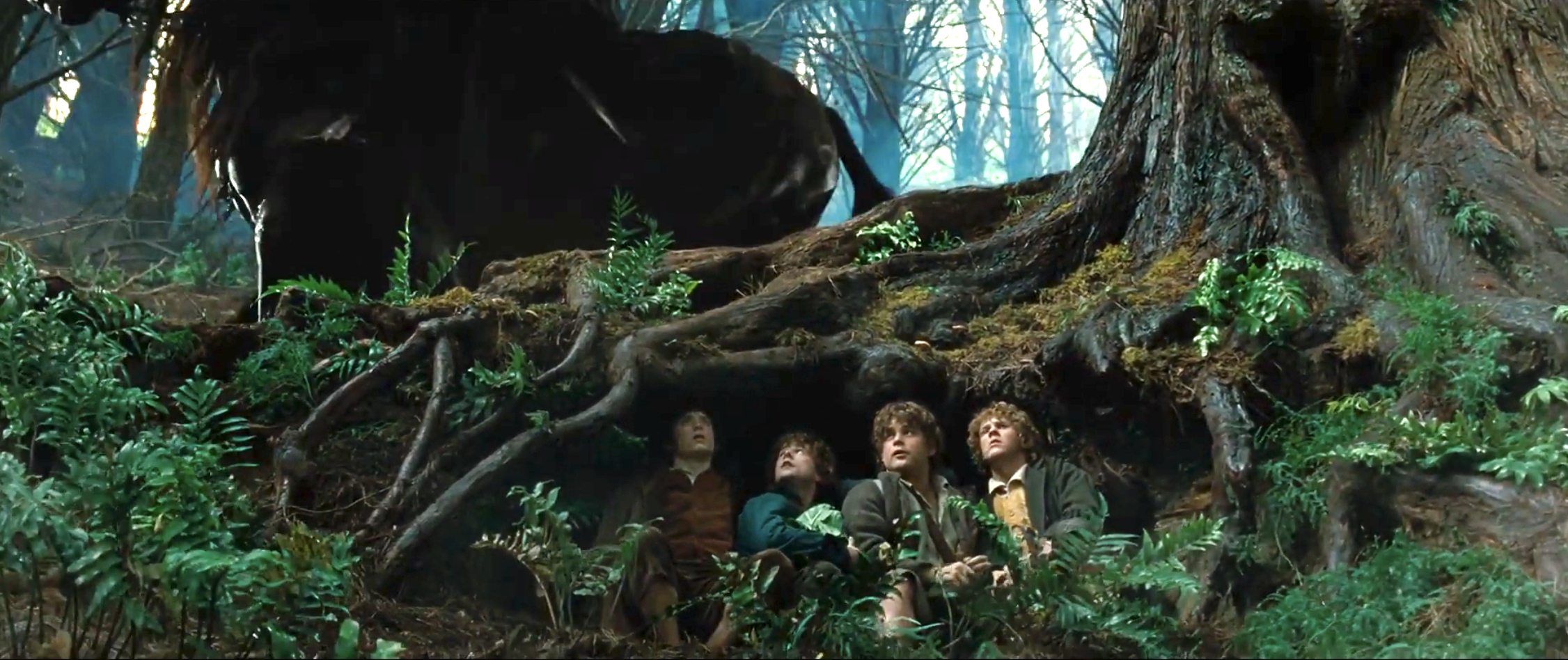 From "Lord of the Rings": Elijah Wood, Sean Astin, Dominic Monaghan, and Billy Boyd hide under tree roots in a forest. The setting appears mystical and dense with foliage