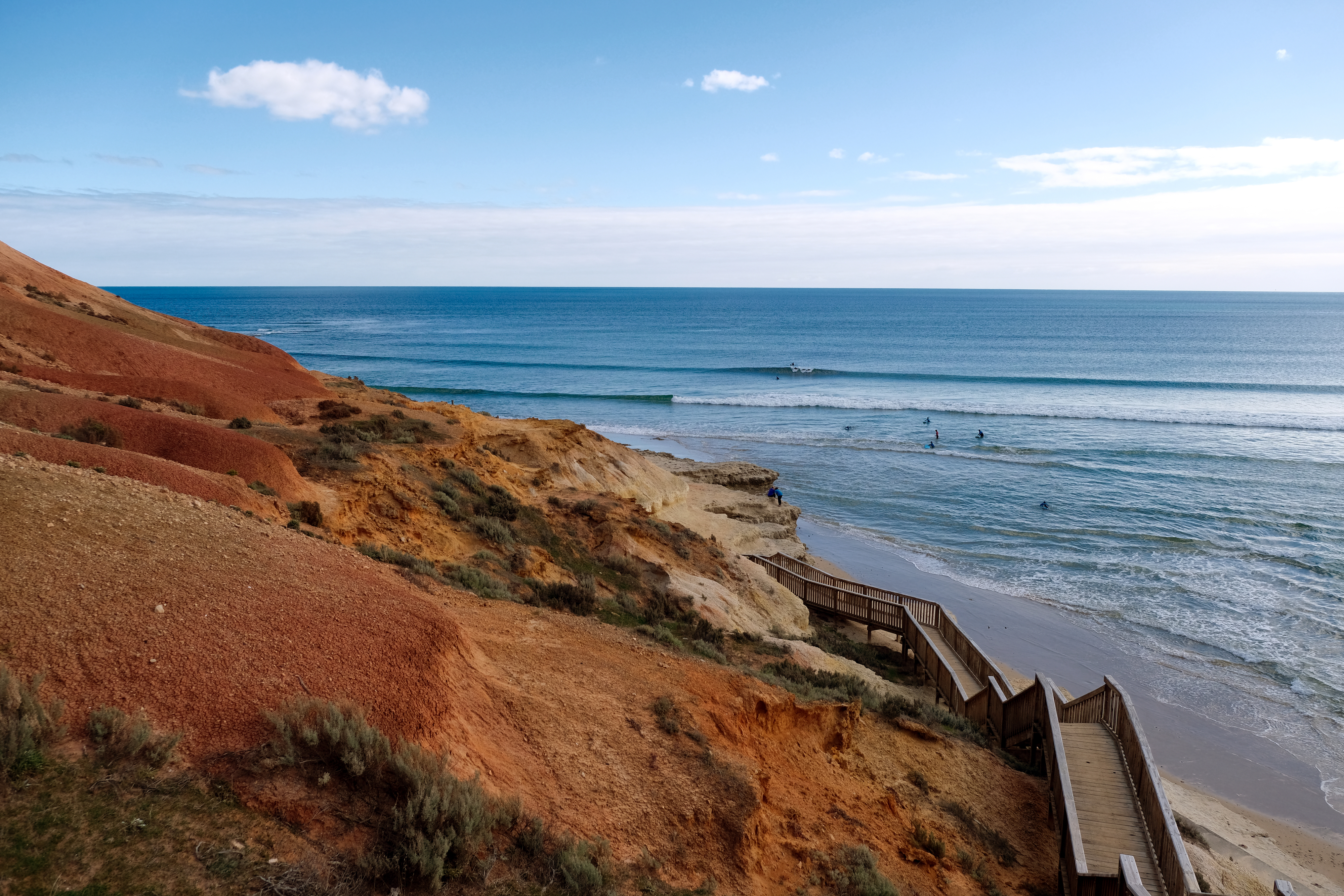 Wooden boardwalk leading down to a sandy beach with surf waves, bordered by rocky cliffs and vegetation under a clear sky