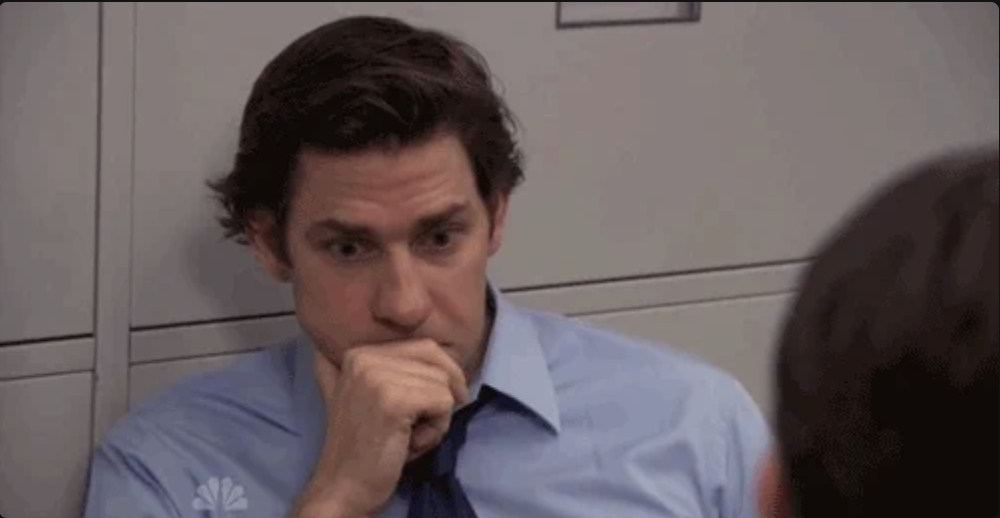 John Krasinski in a thoughtful pose, resting his chin on a clenched fist, from a scene in a workplace setting
