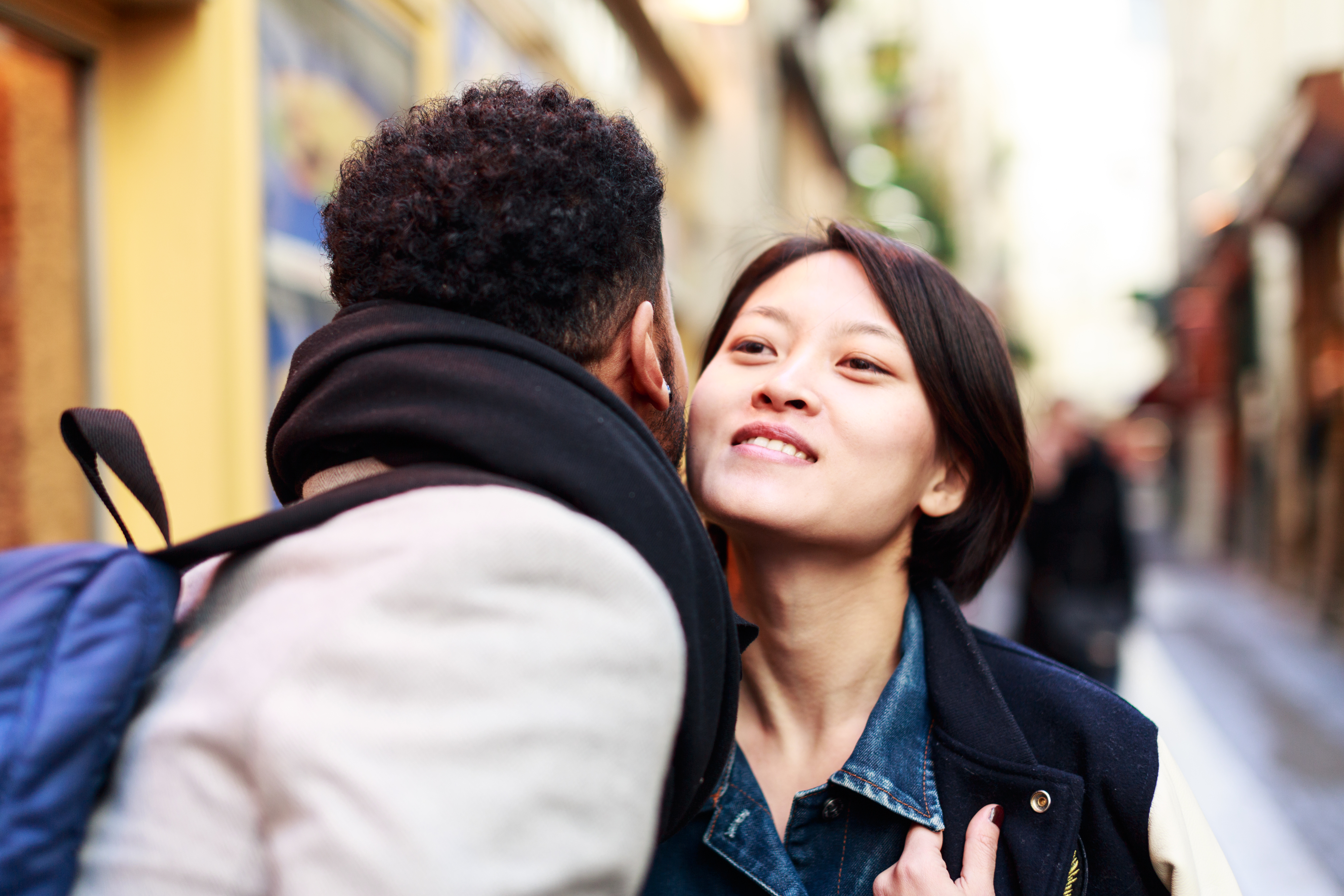A man and a woman, kiss on the cheek on a city street. The man wears a scarf, and the woman wears a denim jacket.