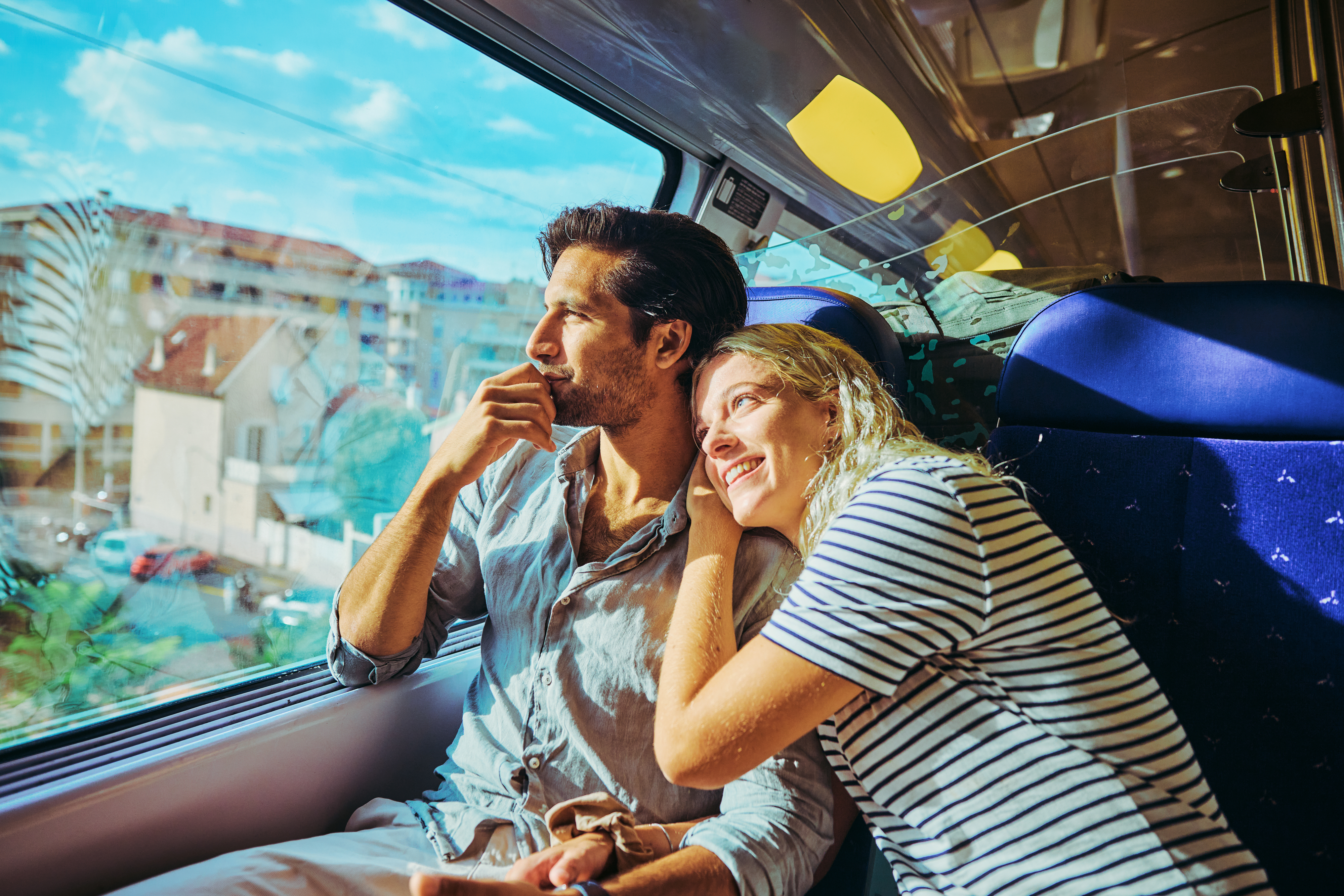 A man and woman sitting on a train, looking out the window. The woman leans affectionately on the man's shoulder. There's a bright scenery outside the window.