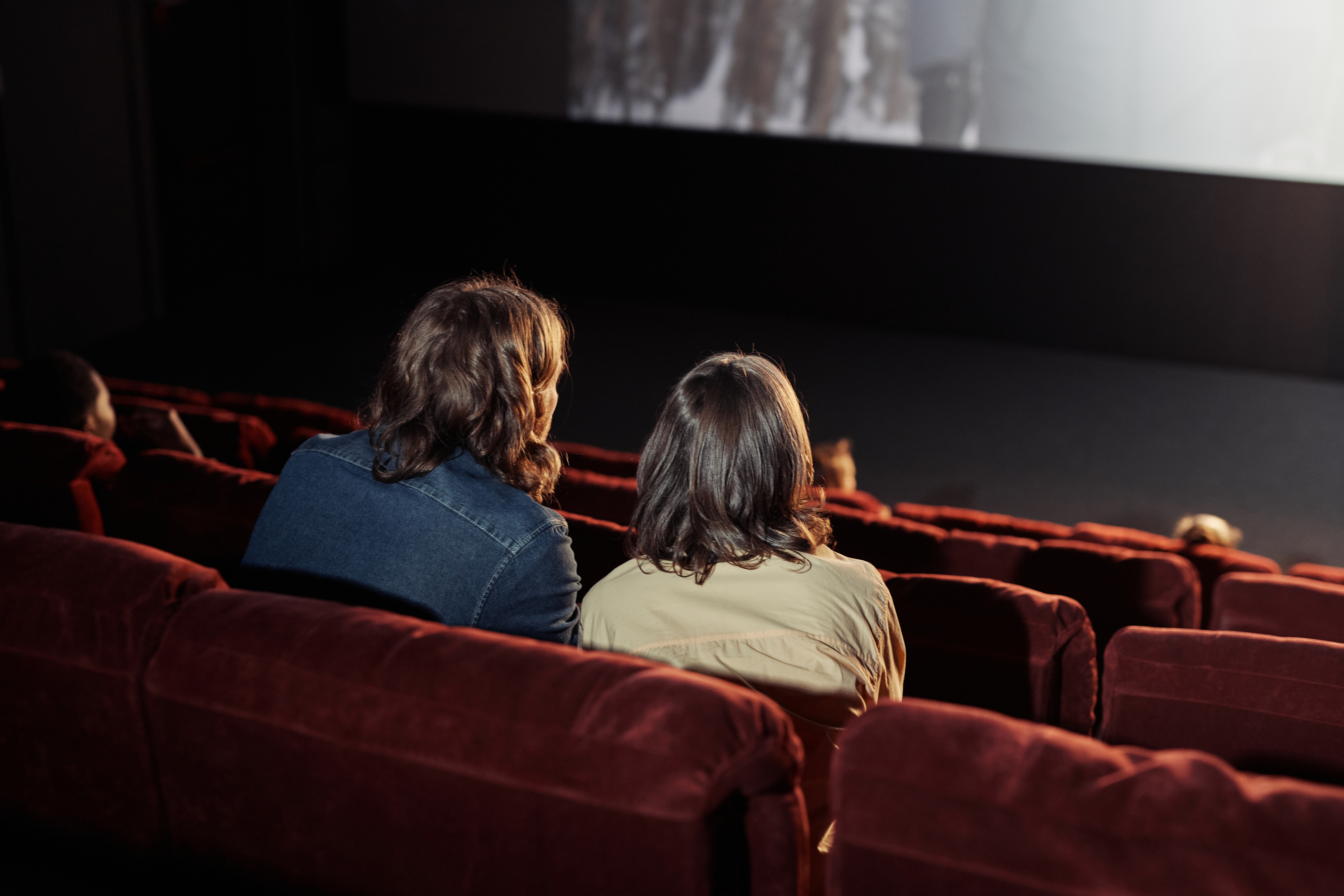 Two people with long hair are sitting in a movie theater, viewing a screen. The theater has red seats