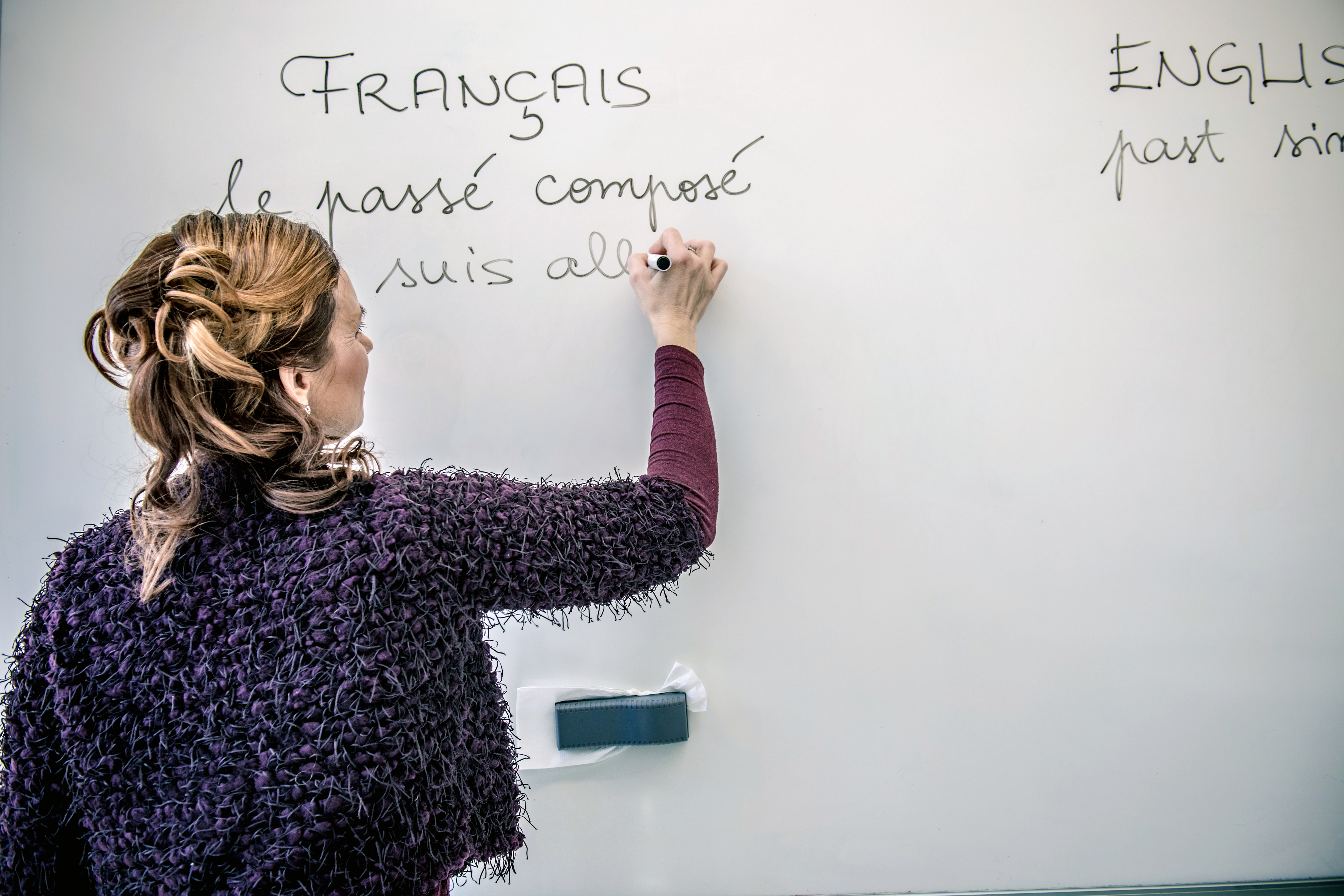 A woman writes French text on a whiteboard. The text reads "FRANÇAIS le passé composé suis all"