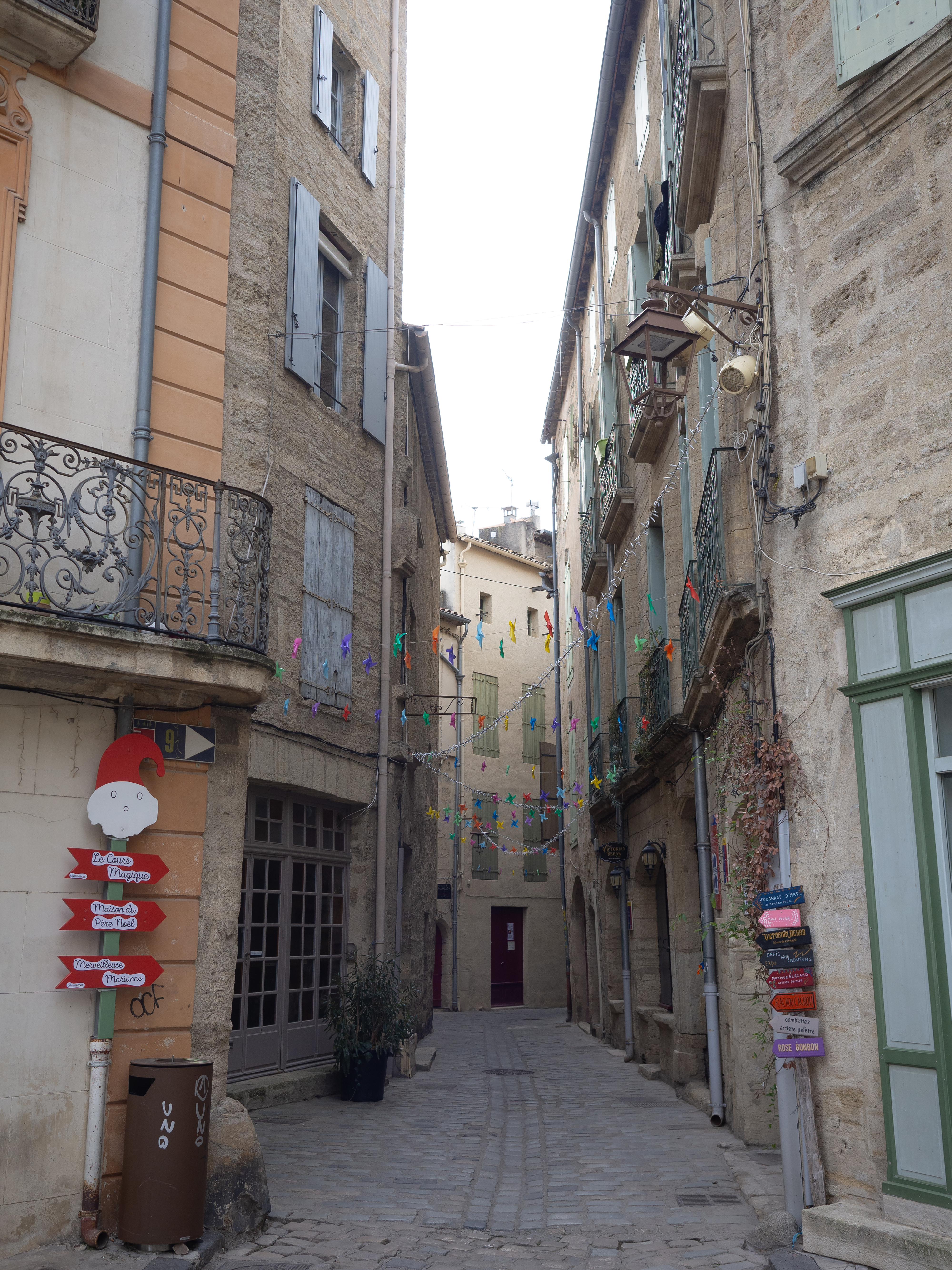 Narrow cobblestone street between tall, old buildings adorned with colorful flags and signs. The street has residential balconies and closed shop fronts.