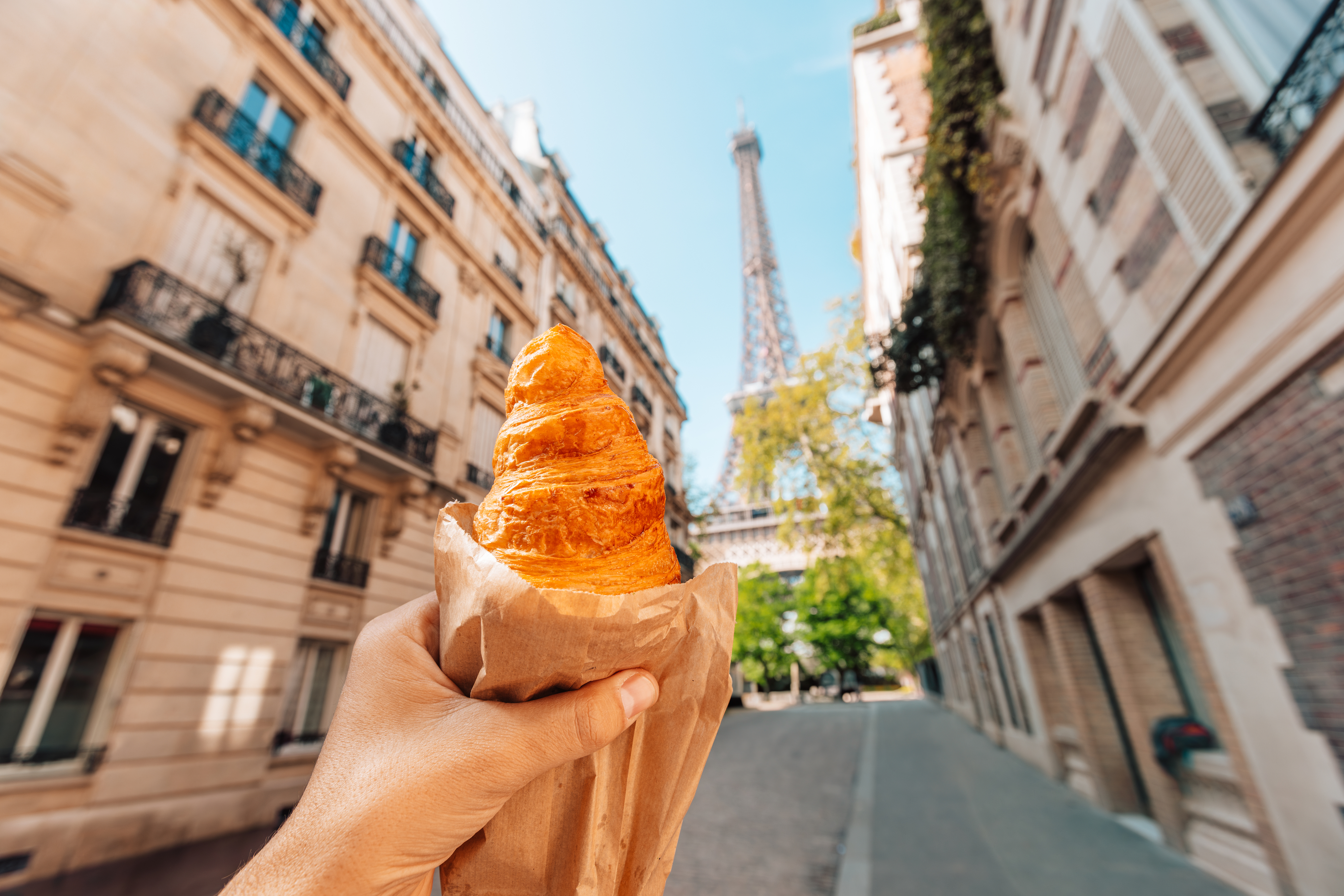 A hand holds a croissant with a street view of Paris and the Eiffel Tower in the background.