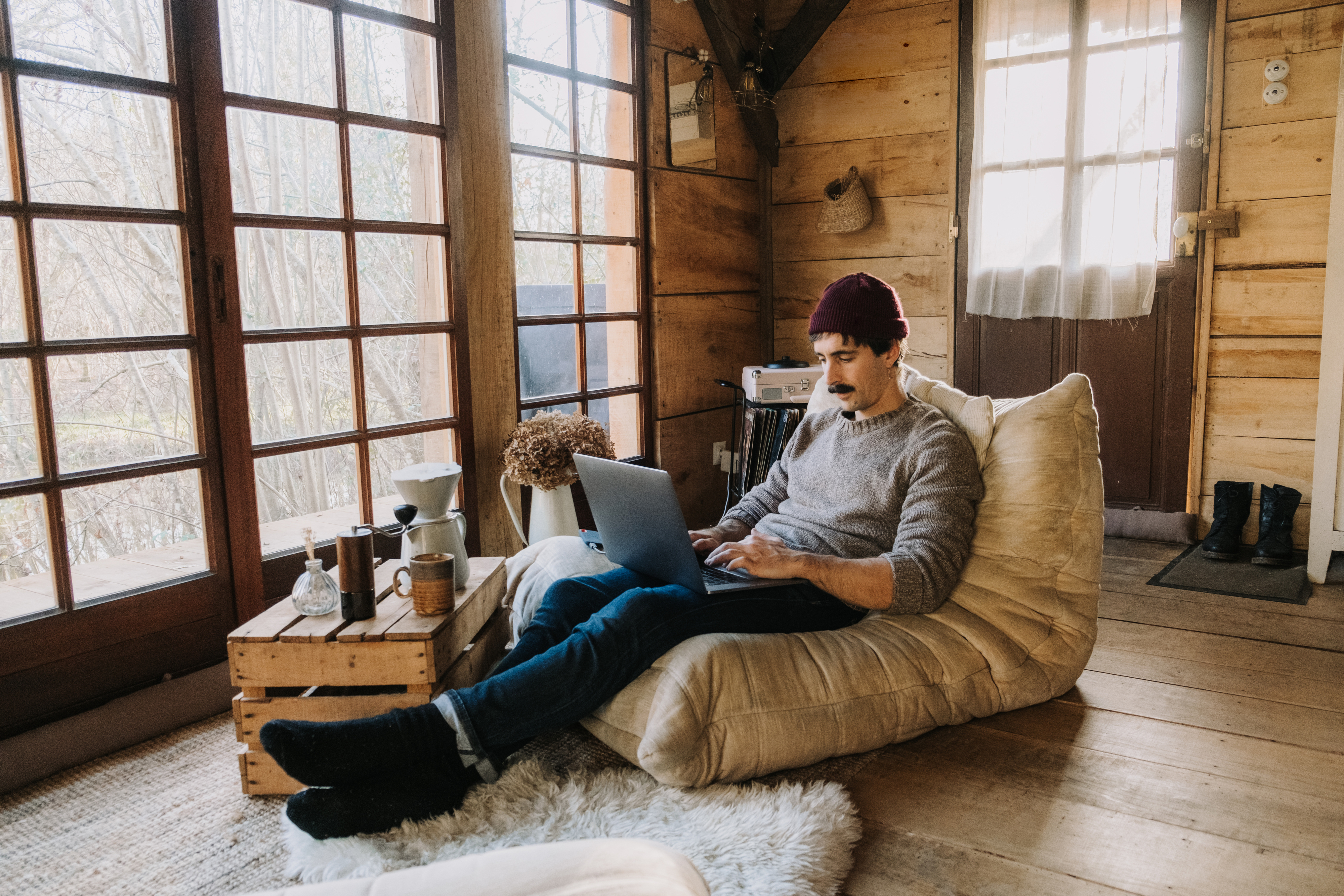 A man sits comfortably on a bean bag chair in a cozy wooden cabin, working on a laptop.