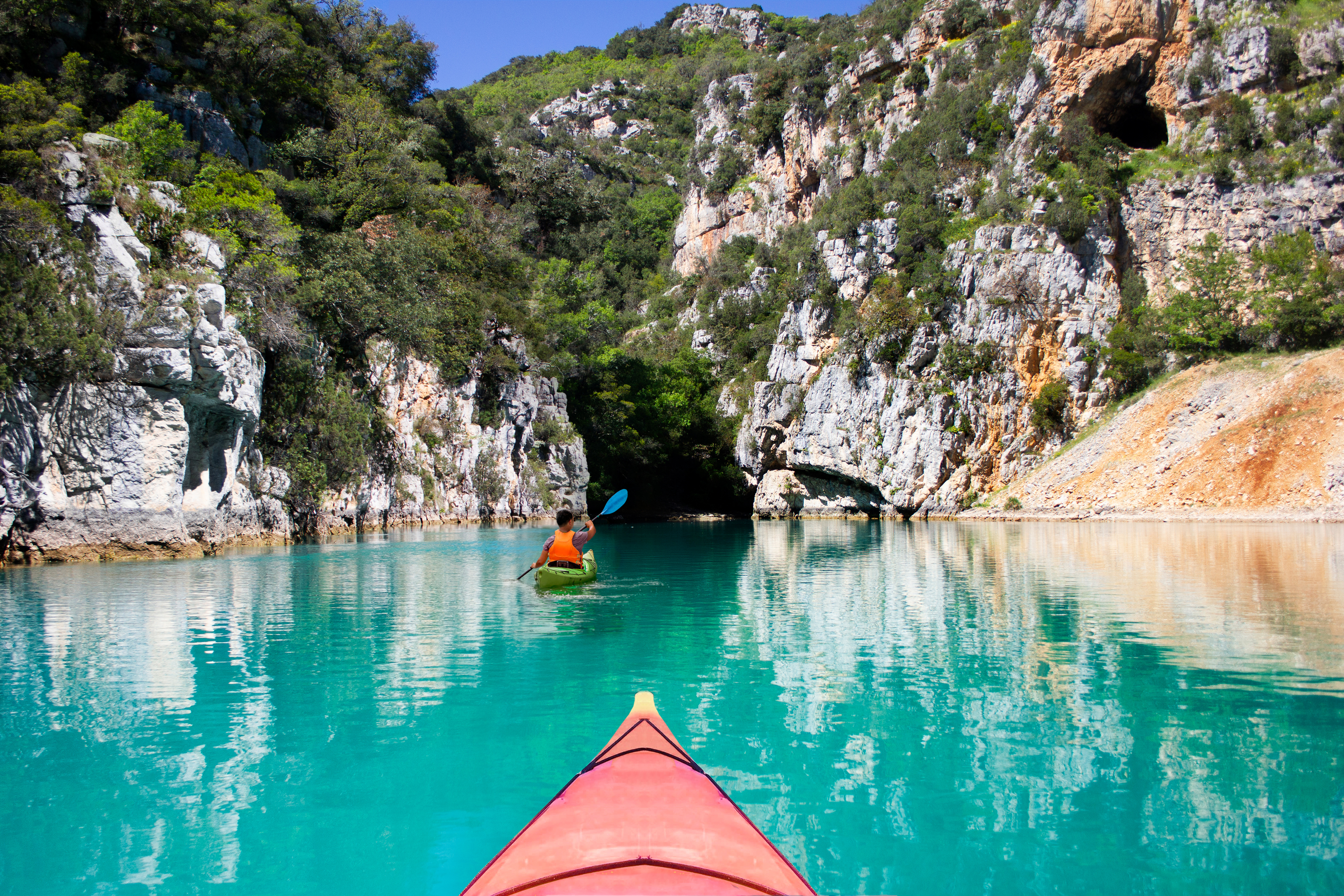 A person in an orange kayak paddles through calm, clear water surrounded by steep rocky cliffs and lush greenery.