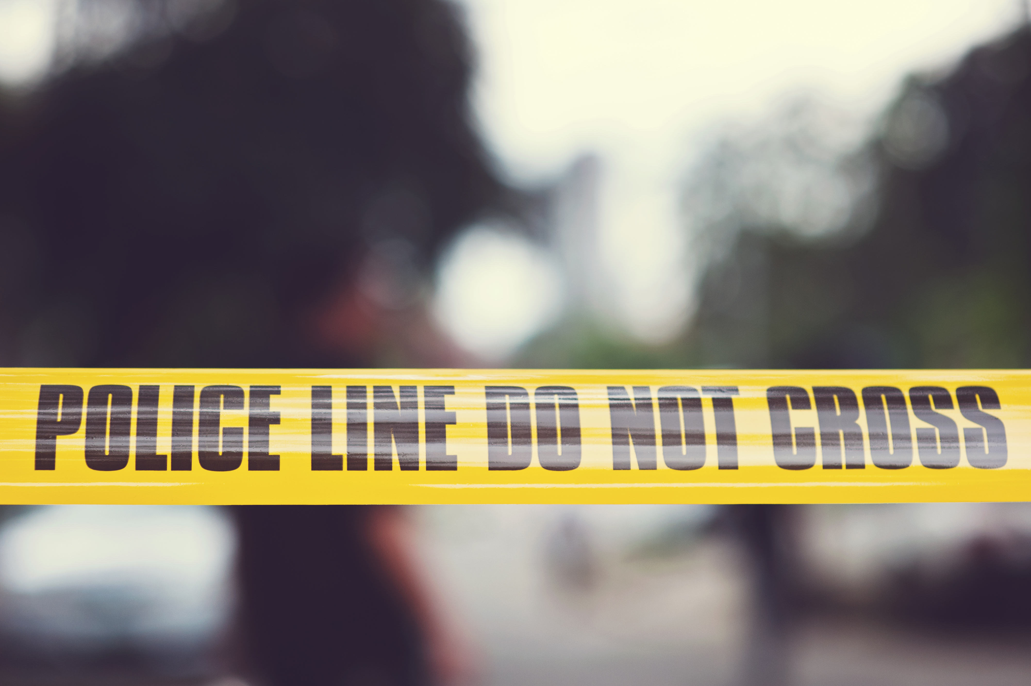 Yellow "Police Line Do Not Cross" tape cordons off an area outside with blurred trees and a building in the background