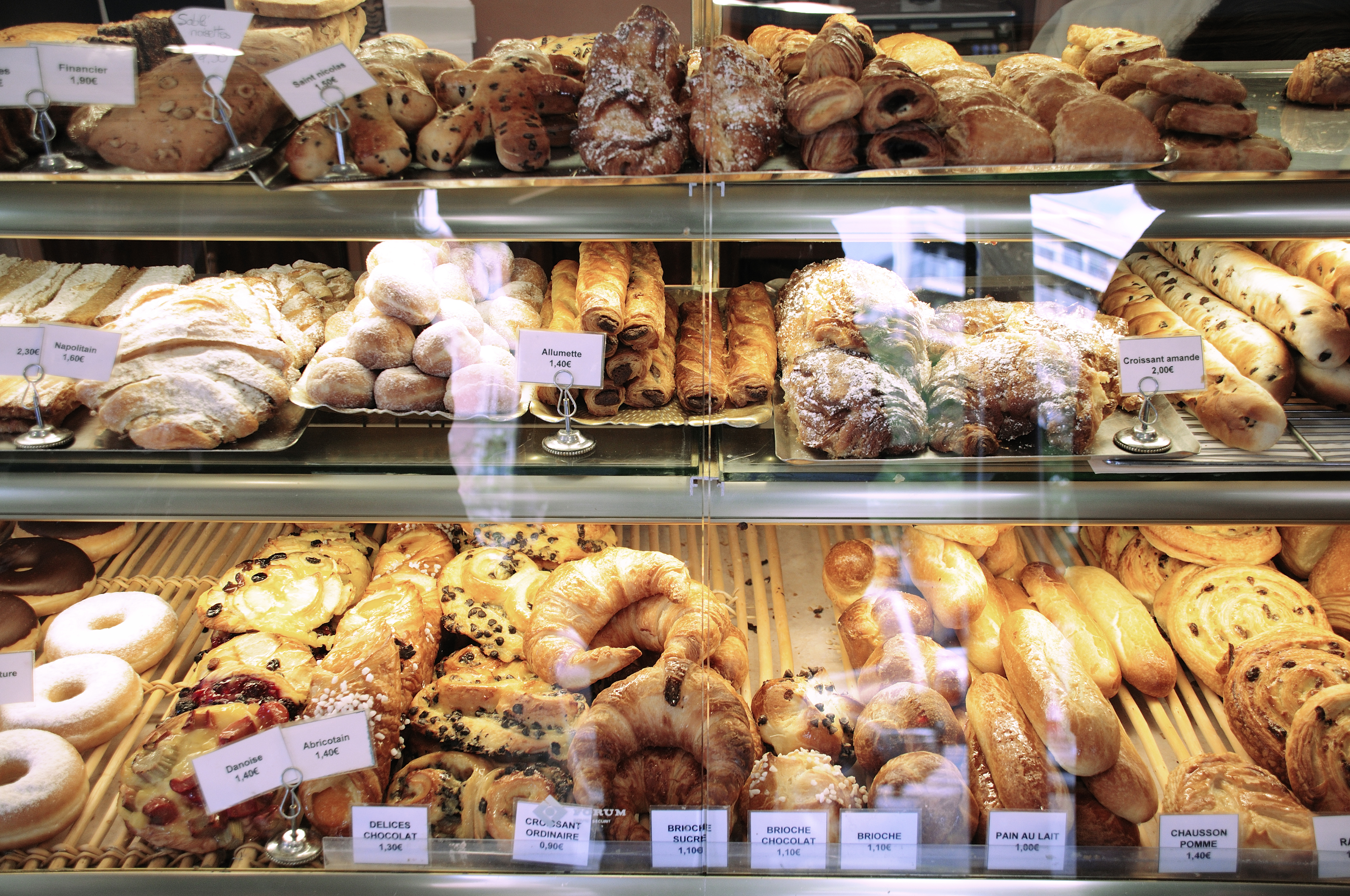 A bakery display case filled with a variety of pastries, such as croissants, donuts, and baguettes. Each item is labeled with its name.