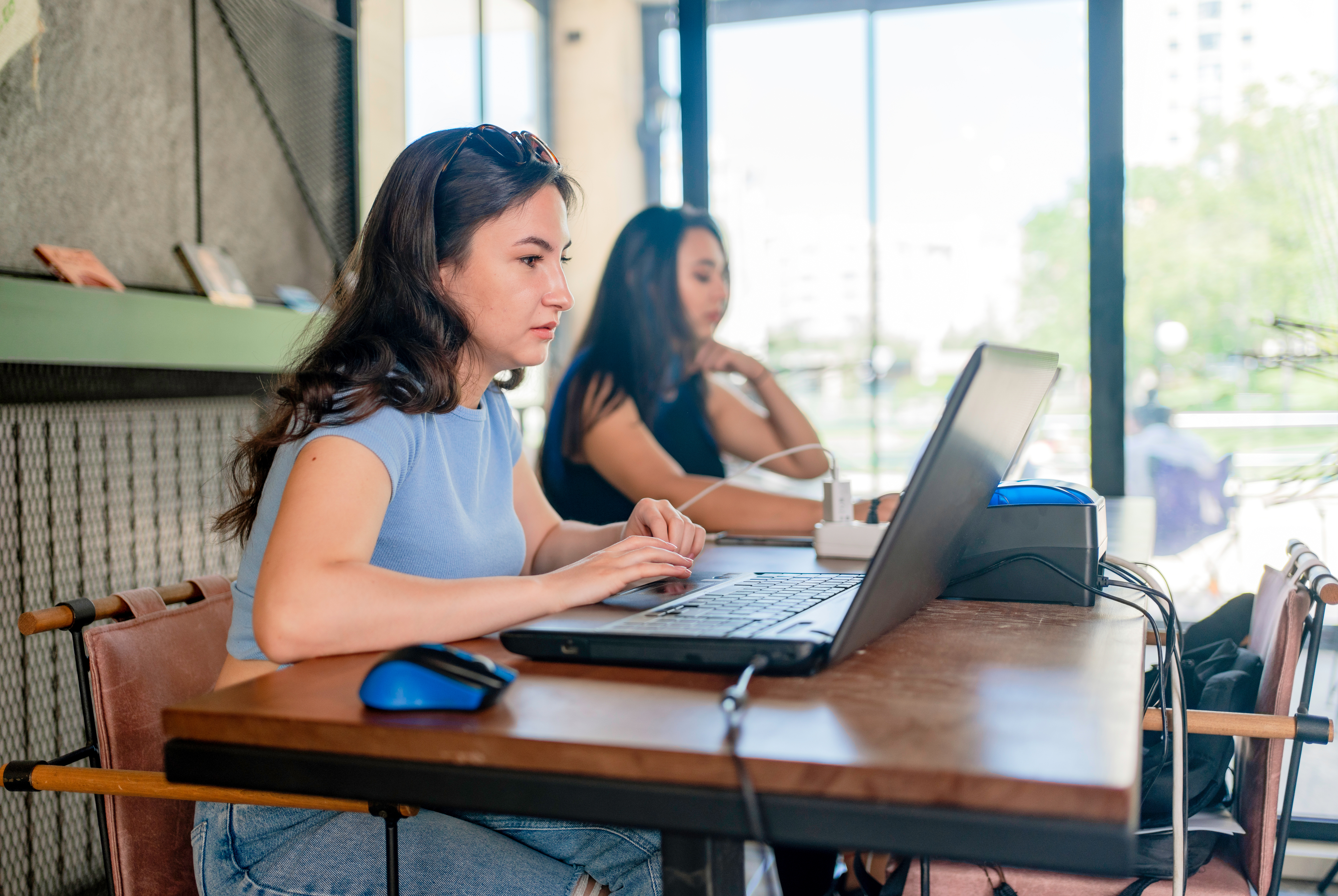 Two women work on laptops at a wooden table in a bright, modern workspace