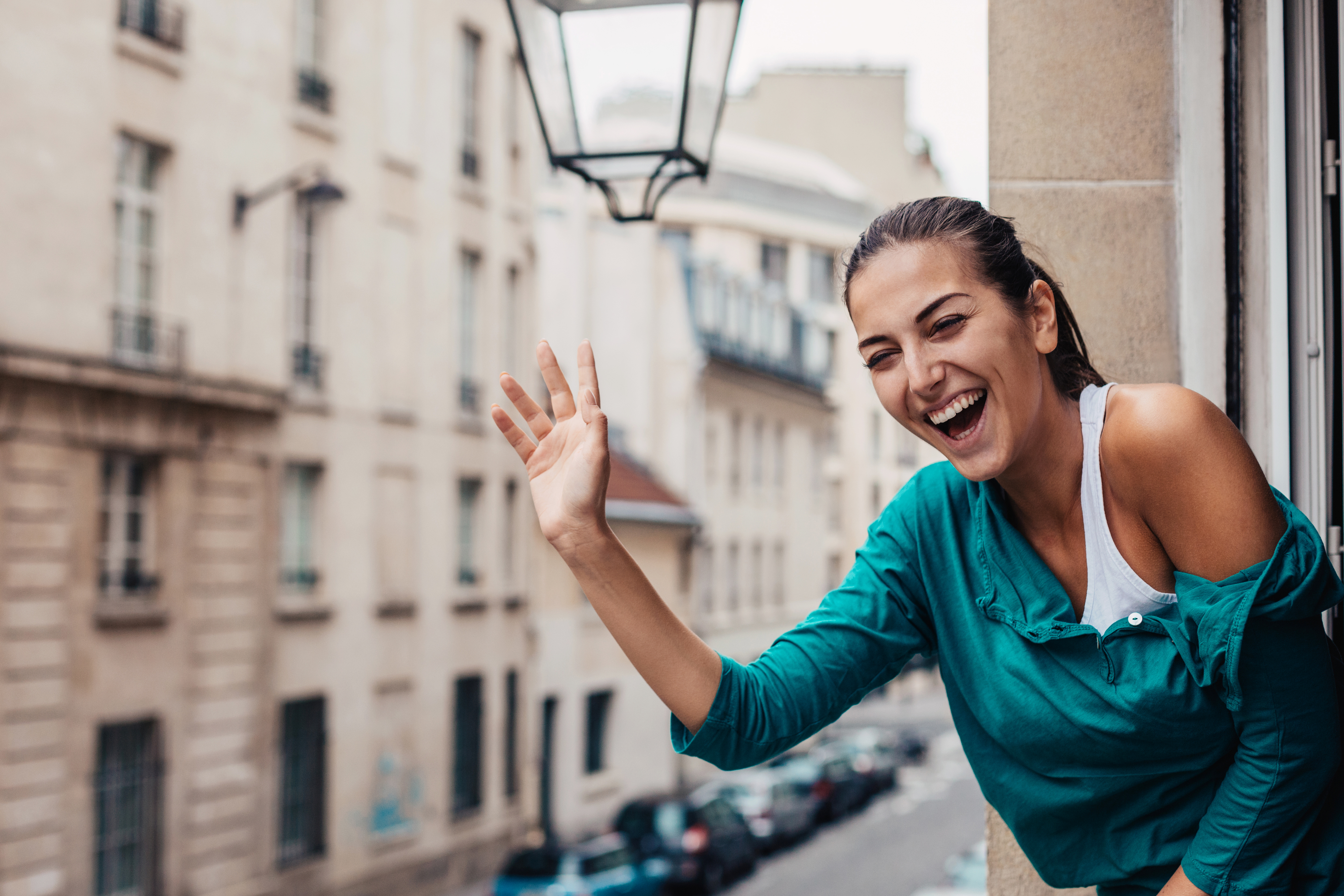 A woman with her hair tied back smiles and waves from an open window of a building, with a street view in the background.