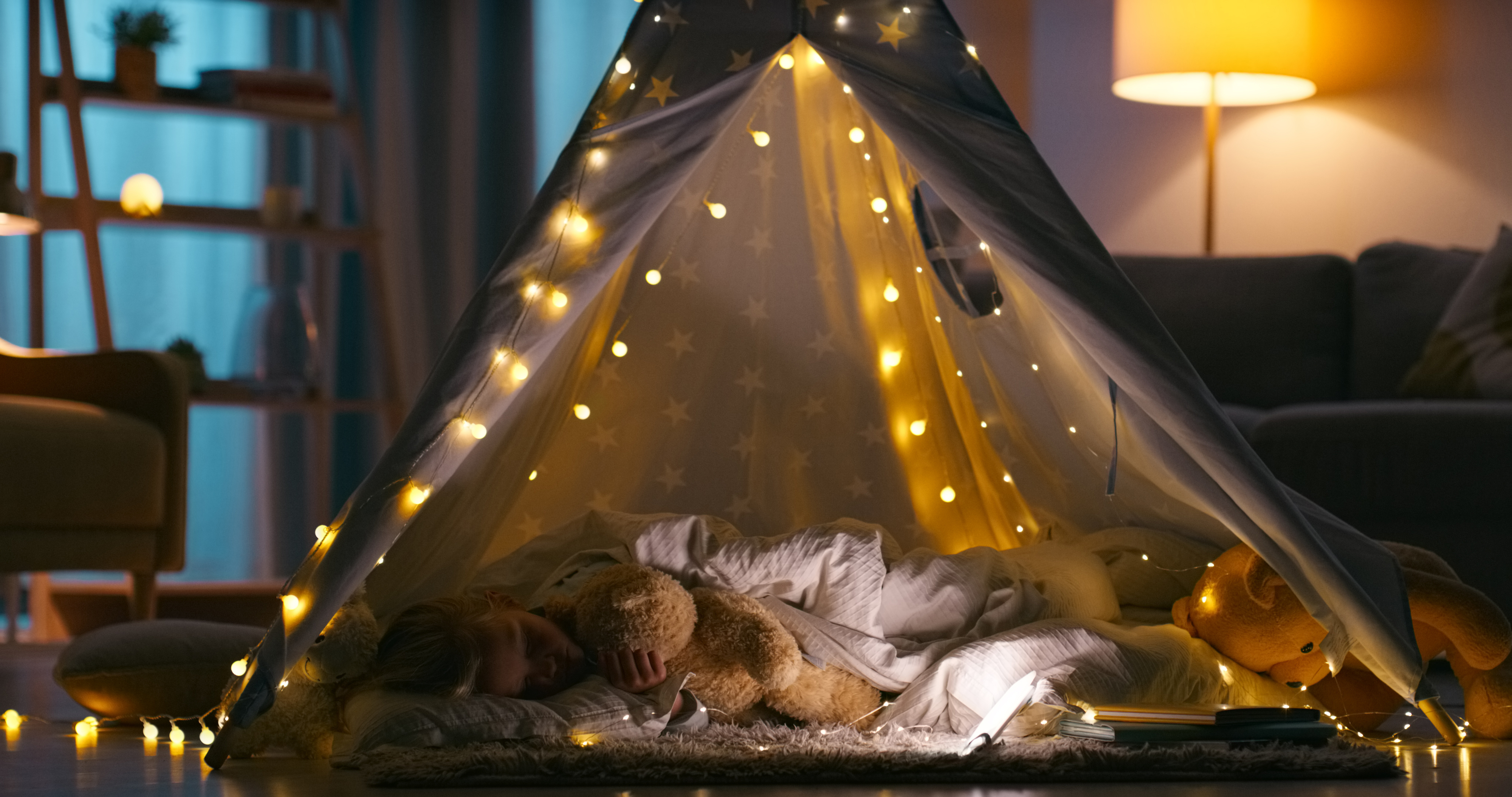Child asleep inside a cozy indoor blanket fort decorated with string lights, holding a teddy bear. The scene is set in a softly lit living room with a lamp in the background