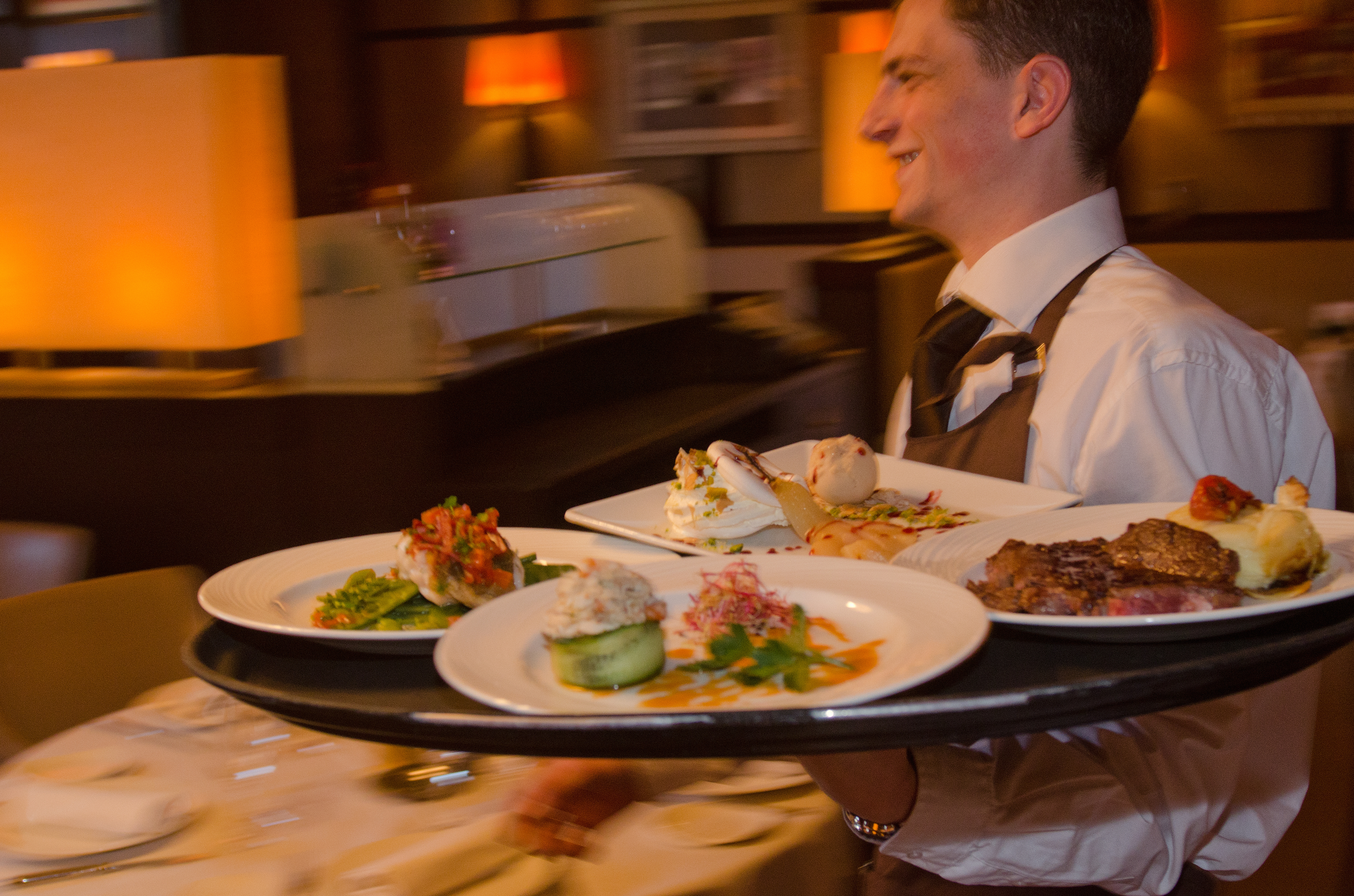A server in a restaurant smiles as he balances a tray with multiple dishes, including steak, seafood, and a dessert.
