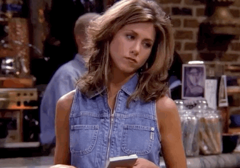 Jennifer Aniston, as Rachel from Friends, stands with a notepad wearing a sleeveless denim shirt in a café setting, looking to the side