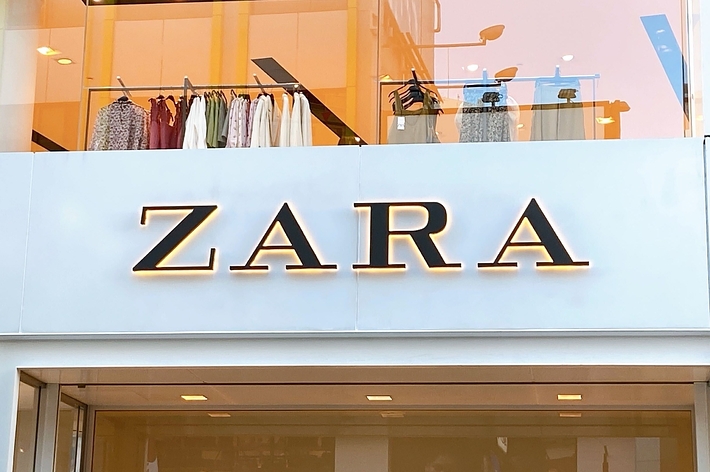 The image shows the front facade of a Zara clothing store, with the company’s name prominently displayed on the sign above the entrance