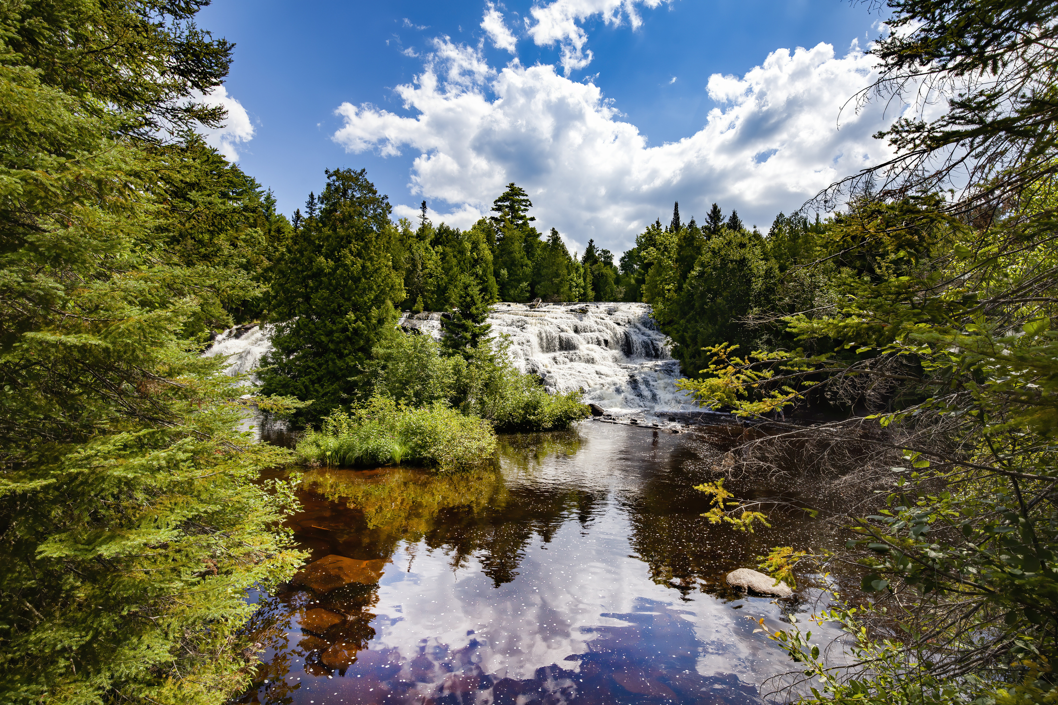 Waterfall in a scenic forest setting, with a river flowing through lush greenery under a bright, cloudy sky