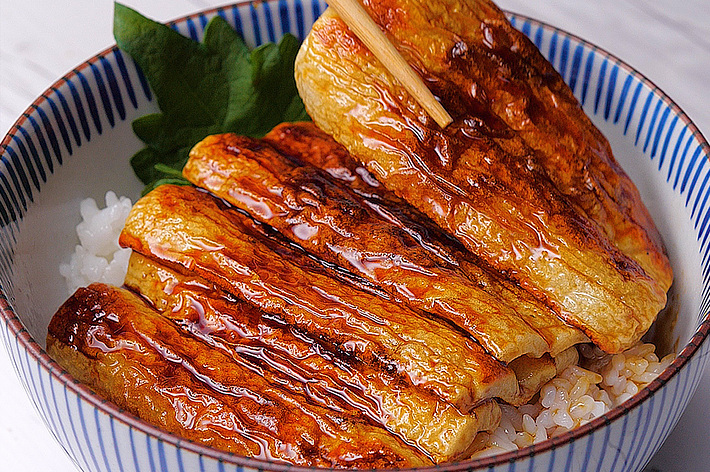 器に盛られたご飯の上に、箸で持ち上げられた蒲焼風の卵が並んでいる。