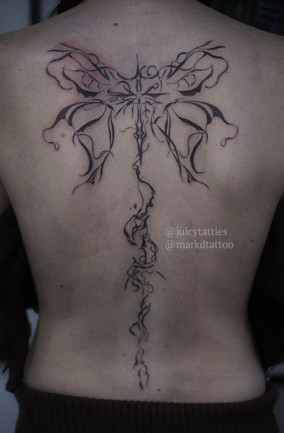 A photo of a person's back with a detailed tattoo of intricate, symmetrical designs resembling wings and flowing lines down the spine. Text reads "@juicytatties @markdtattoo"