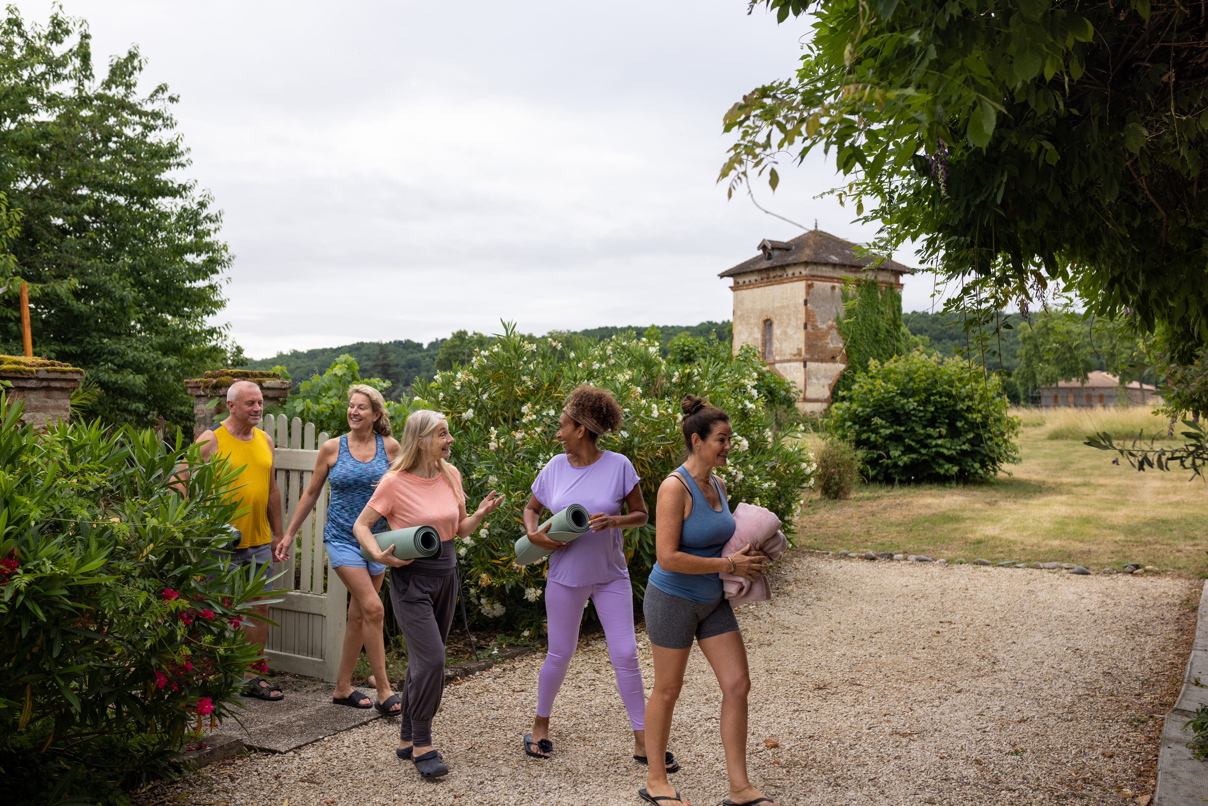 A group of five adults carrying yoga mats, walking through a garden pathway, smiling and talking to each other. A building and greenery is in the background.
