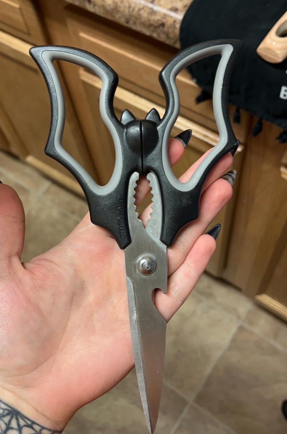 A hand holds a pair of black kitchen shears with a built-in bottle opener near a kitchen sink