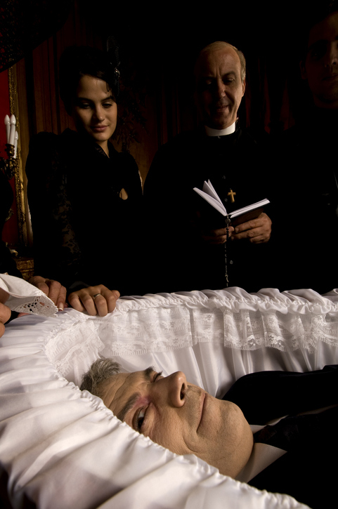 A woman and a priest stand beside an elderly man lying in a white-lined coffin, appearing to participate in a funeral service