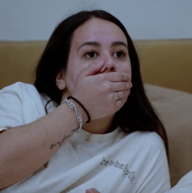 A woman sits on a couch, covering her mouth with her hand, wearing a plain T-shirt with a bracelet and tattoo visible on her arm