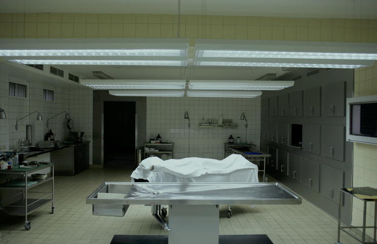 A hospital morgue room with steel tables, a body covered with a white sheet, and medical equipment on wheeled carts