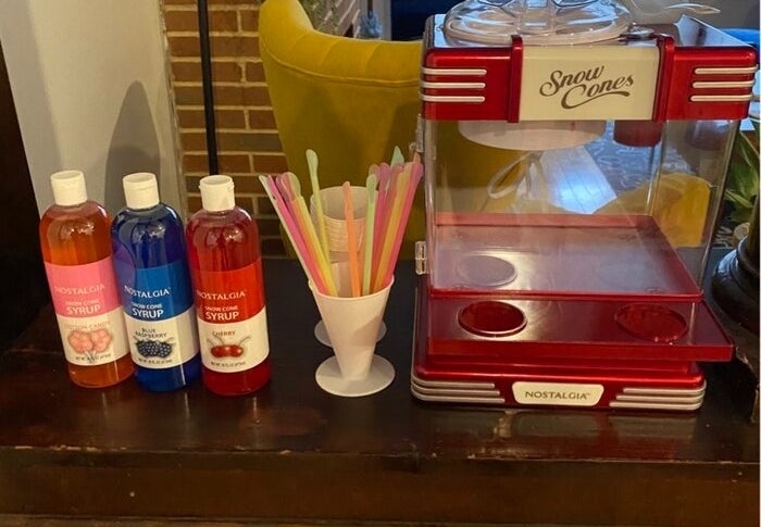 Snow cone machine and assorted syrups (cherry, blue raspberry, grape) with plastic spoon straws displayed on a counter in a home setting