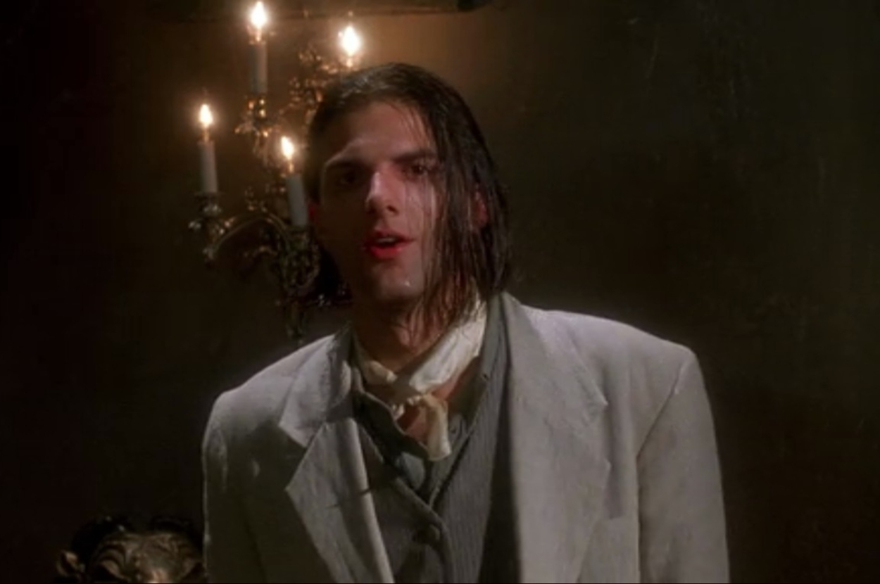A man with wet slicked-back hair wears a light-colored suit with a cravat, standing in front of a candelabra in a dark room
